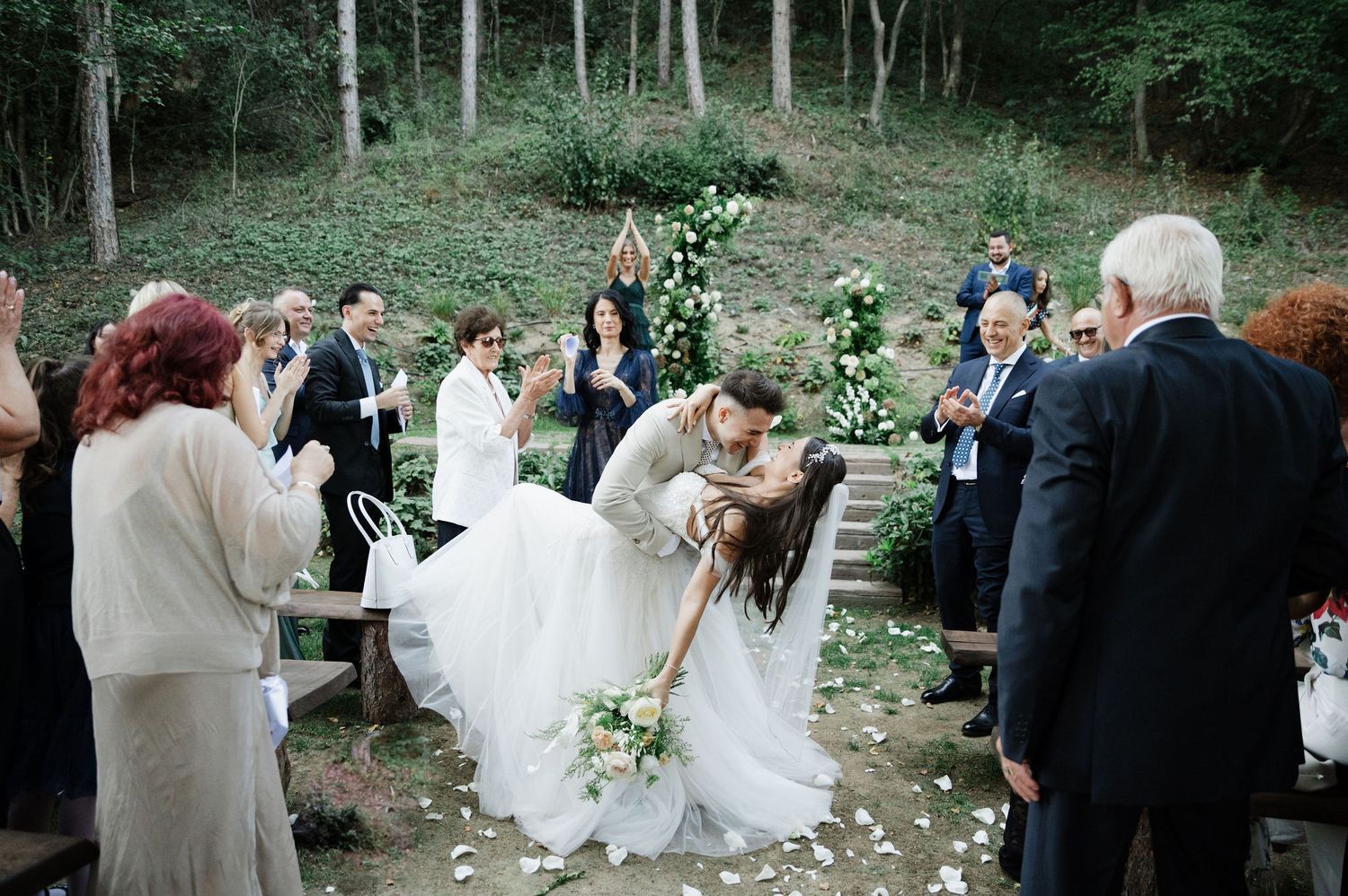 Wedding guests celebrate in a wooded outdoor venue decorated with white flowers and greenery.