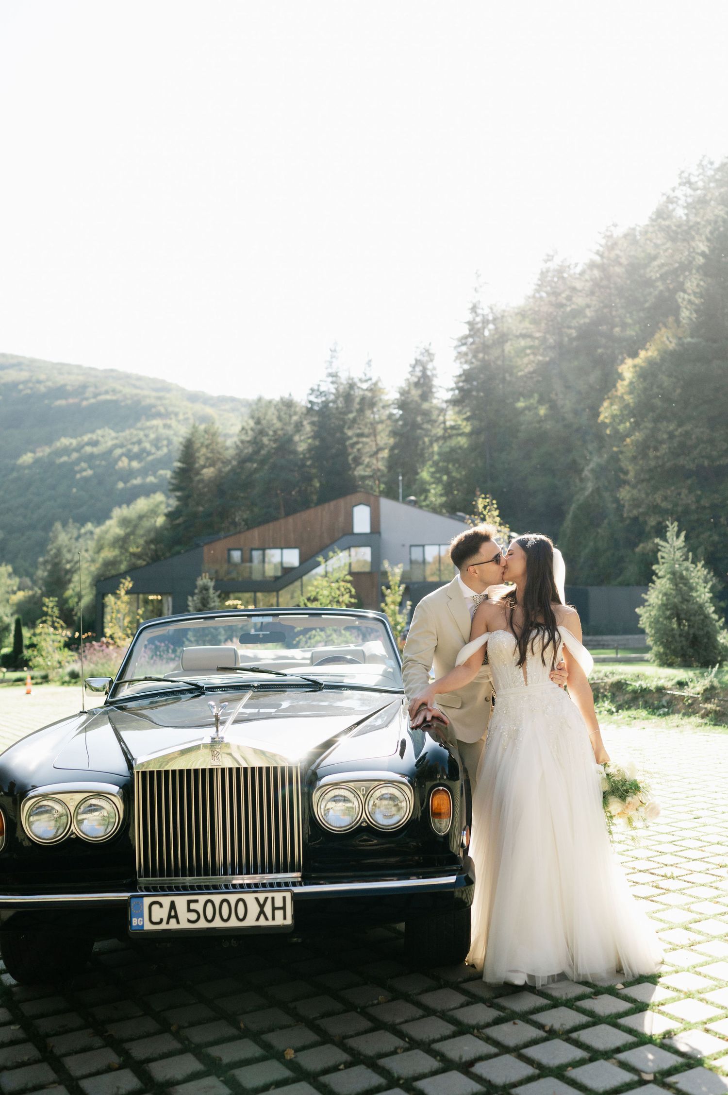 A classic Rolls Royce serves as backdrop for a romantic wedding moment against a mountain landscape.