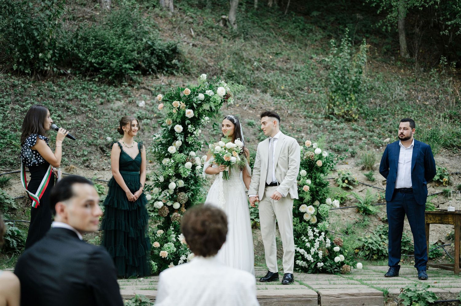 Outdoor wedding ceremony in a wooded setting with stone steps and white floral arrangements.