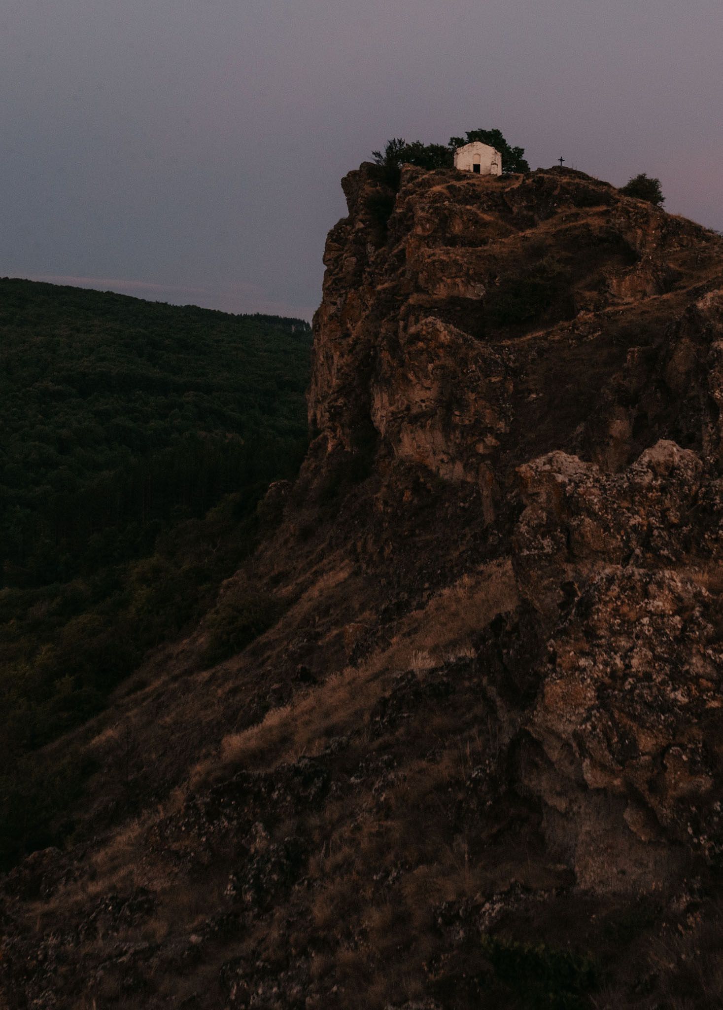 Rocky mountain cliffs silhouetted against a purple dusk sky with a small structure at the peak.