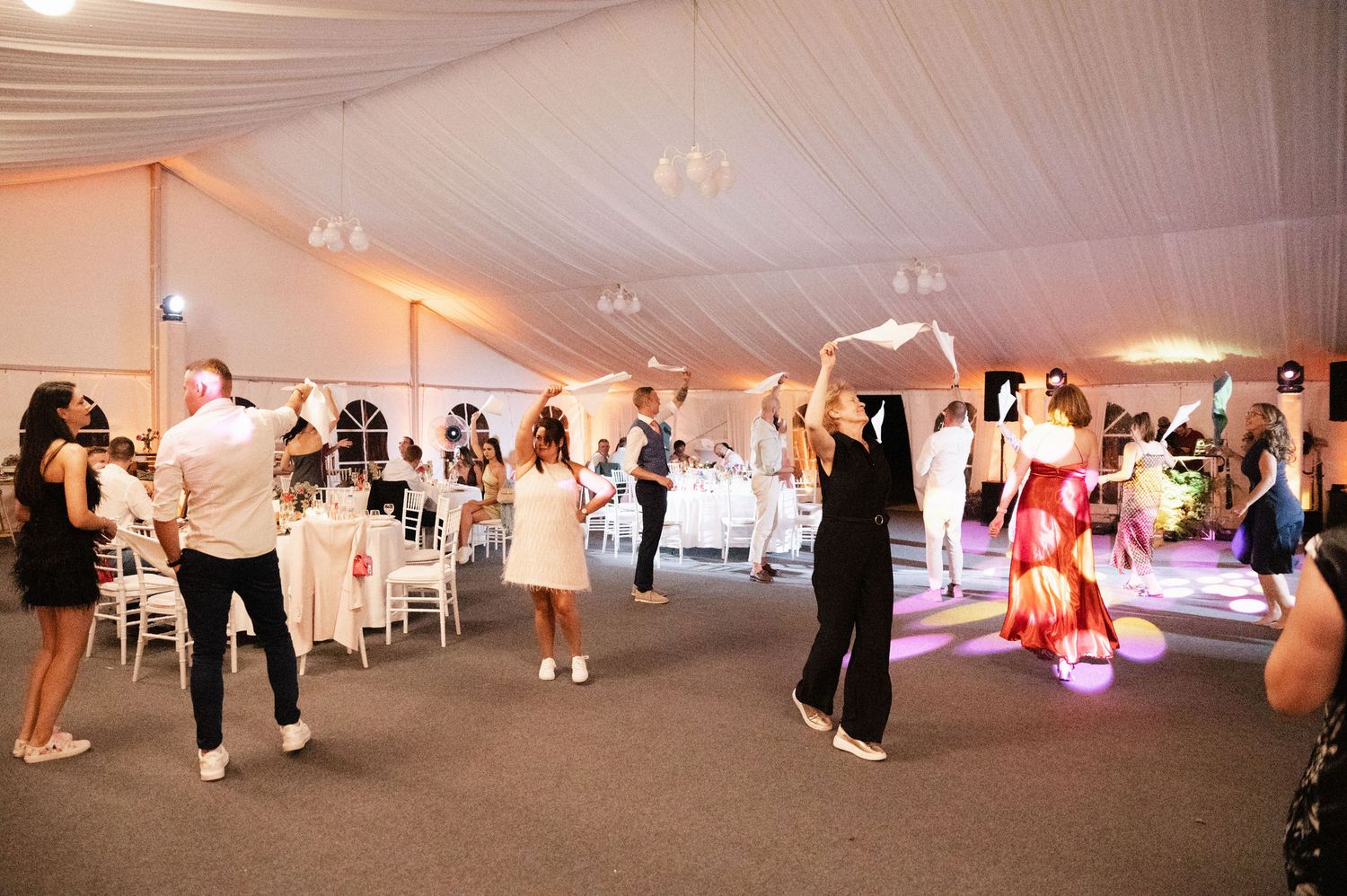 Wedding guests dance and celebrate under a white tent at an evening reception with colorful uplighting.
