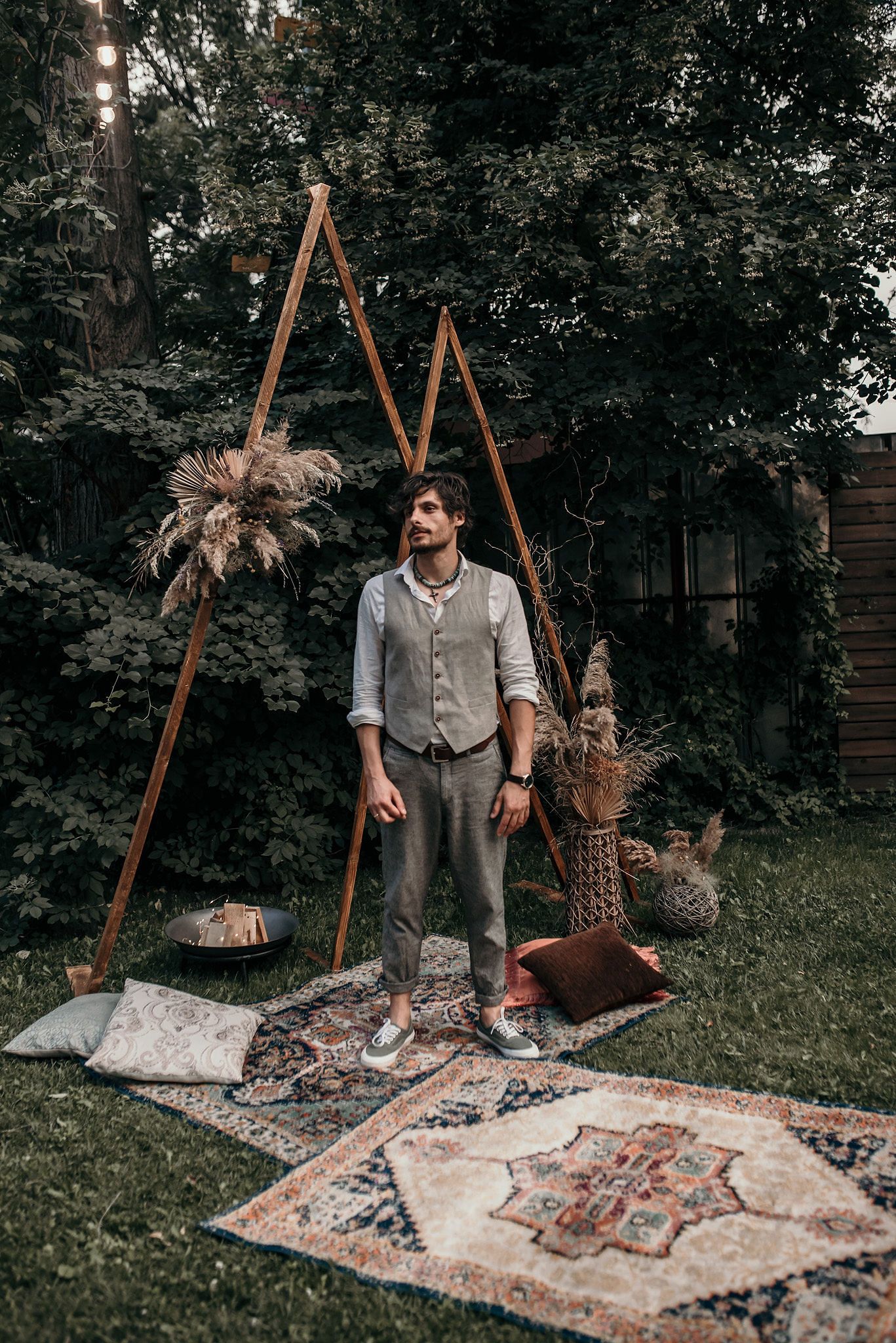 Outdoor scene with decorative wooden tripods and vintage rugs laid out on grass.