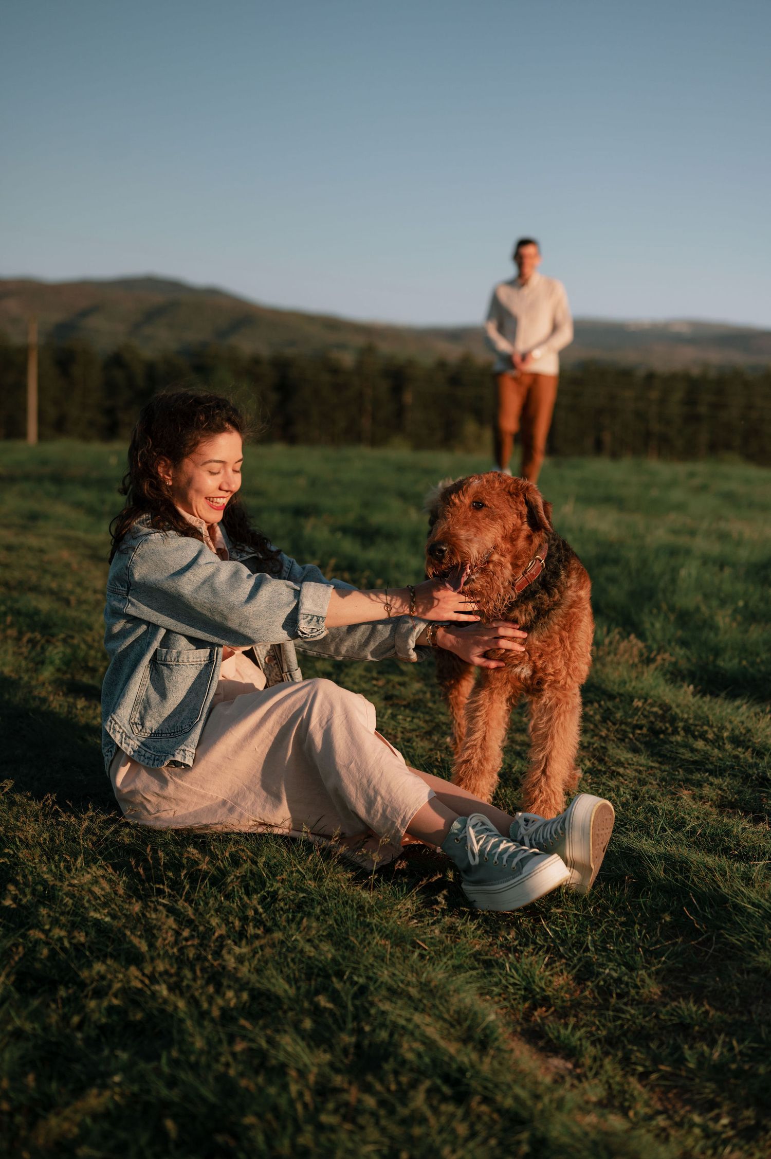 Person petting a brown dog on grassy hillside while another stands in background.