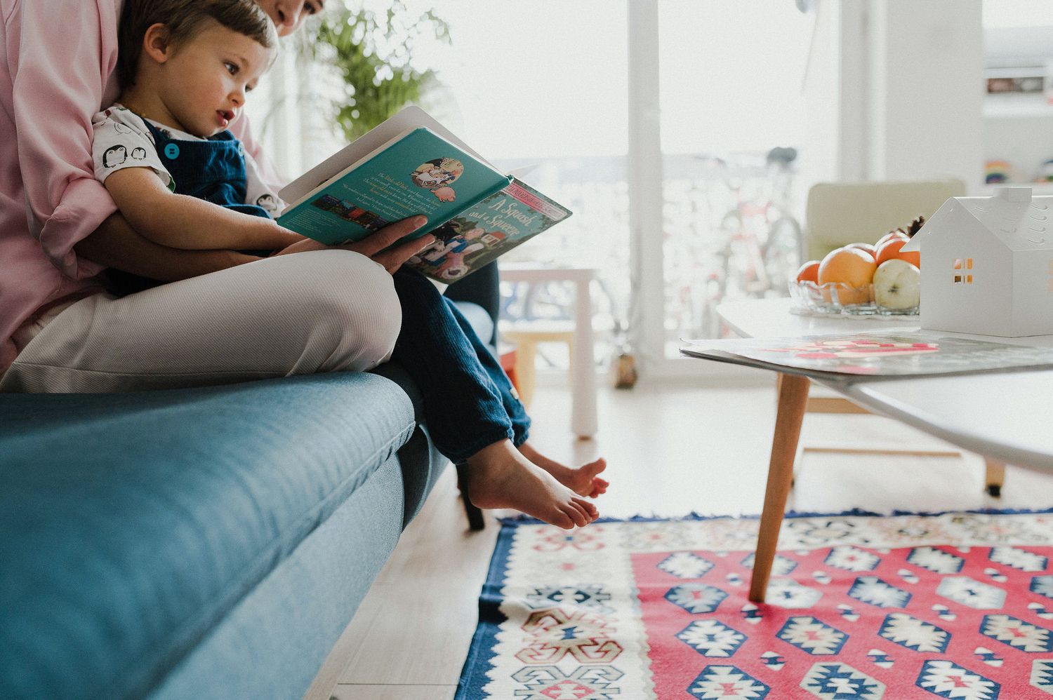 Child sits on couch reading colorful storybook in bright living room with patterned rug.