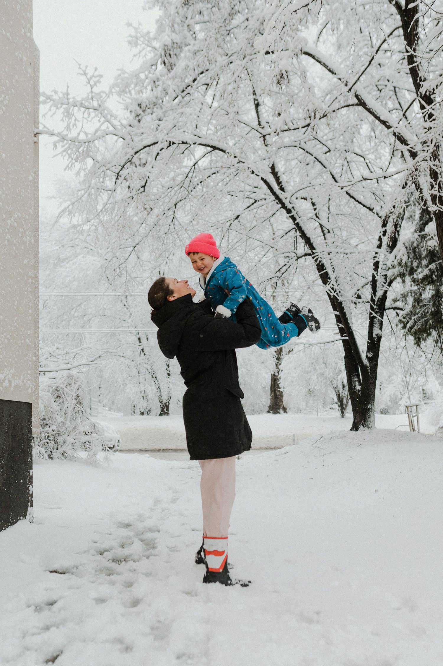 A parent lifts a small child in a blue coat against a snowy winter landscape with frost-covered trees.