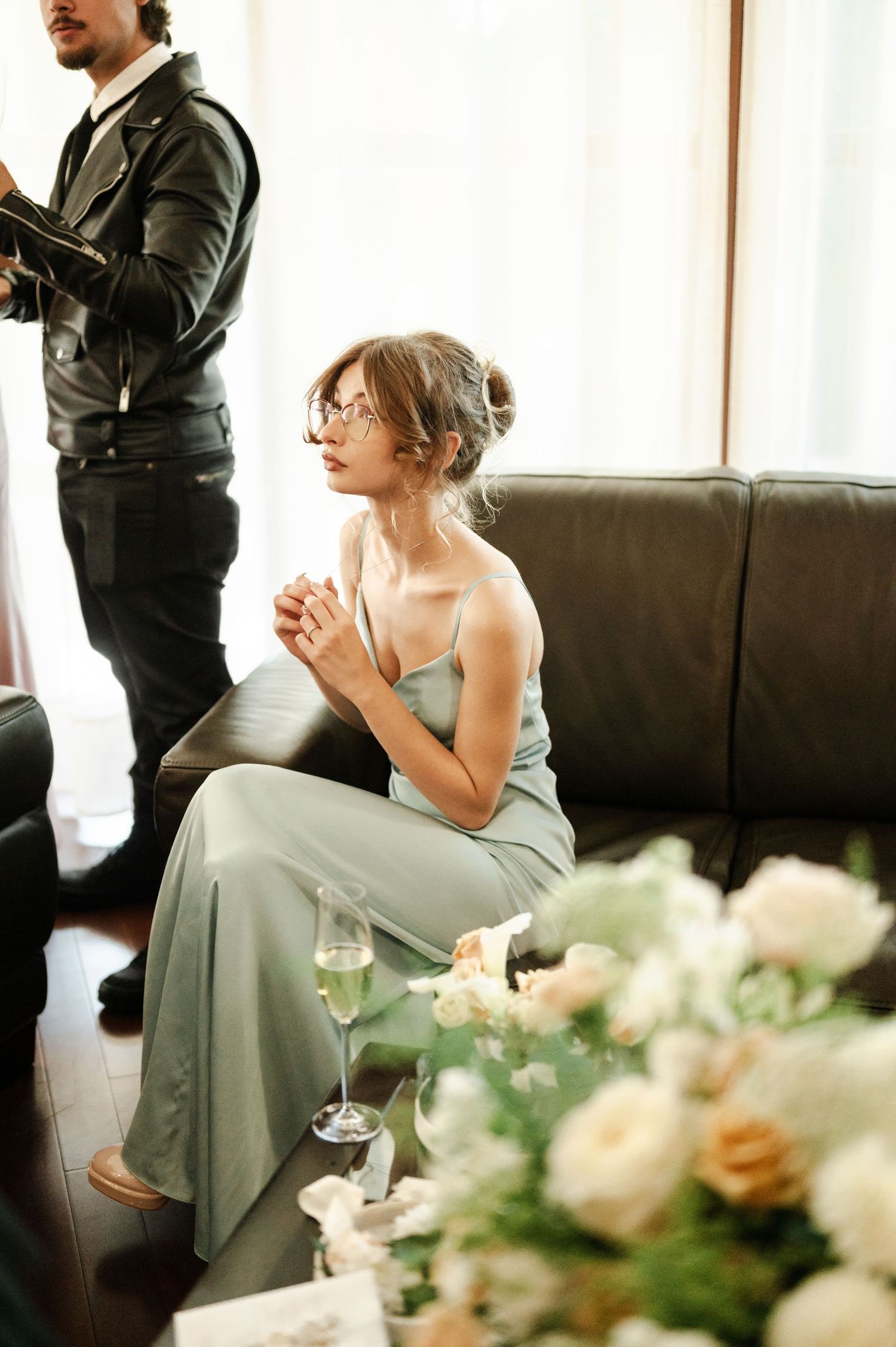 A woman in a sage green dress sits near floral centerpieces at an event.
