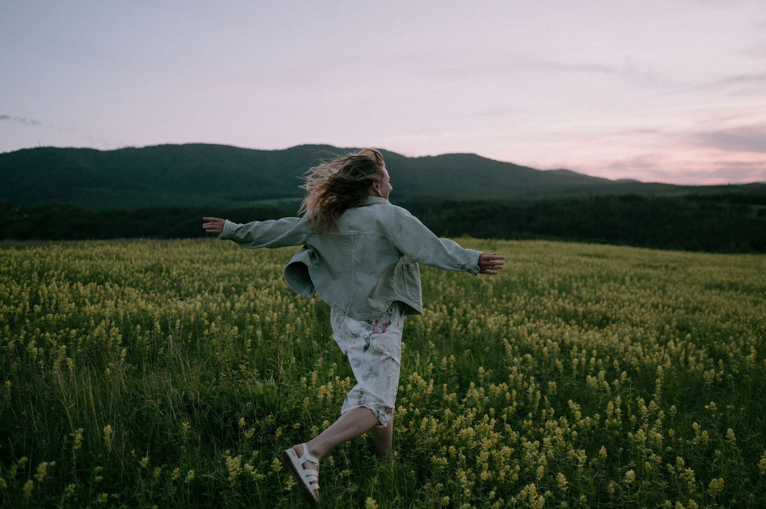 A couple runs playfully through a blooming yellow flower field at sunset with mountains in the background.