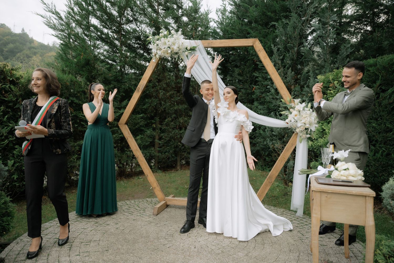 A celebratory moment at an outdoor wedding ceremony with a hexagonal wooden arch decorated with floral arrangements.