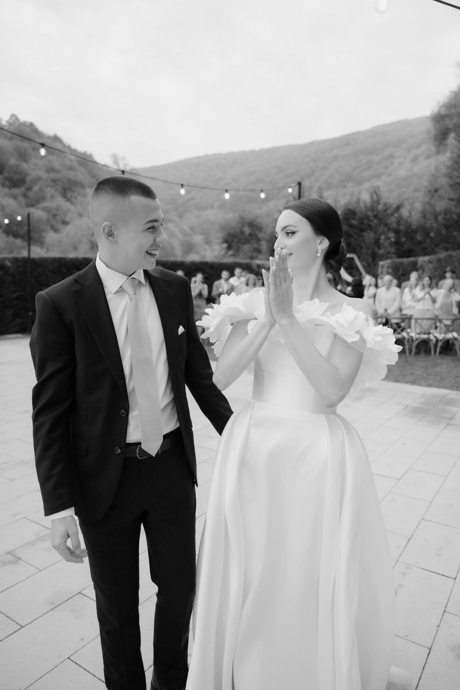 Black and white wedding photo of a couple sharing a candid moment on a stone patio with string lights and hills in background.