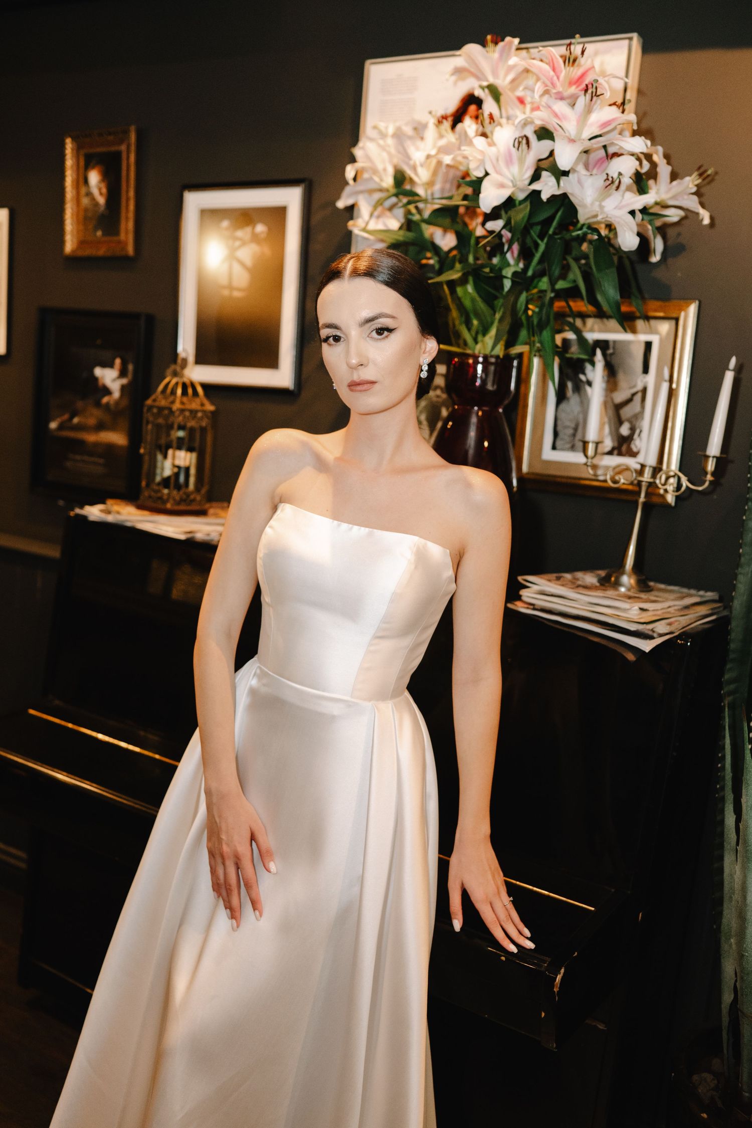 A bride in a classic white satin wedding gown poses elegantly in a dimly lit room with floral arrangements.
