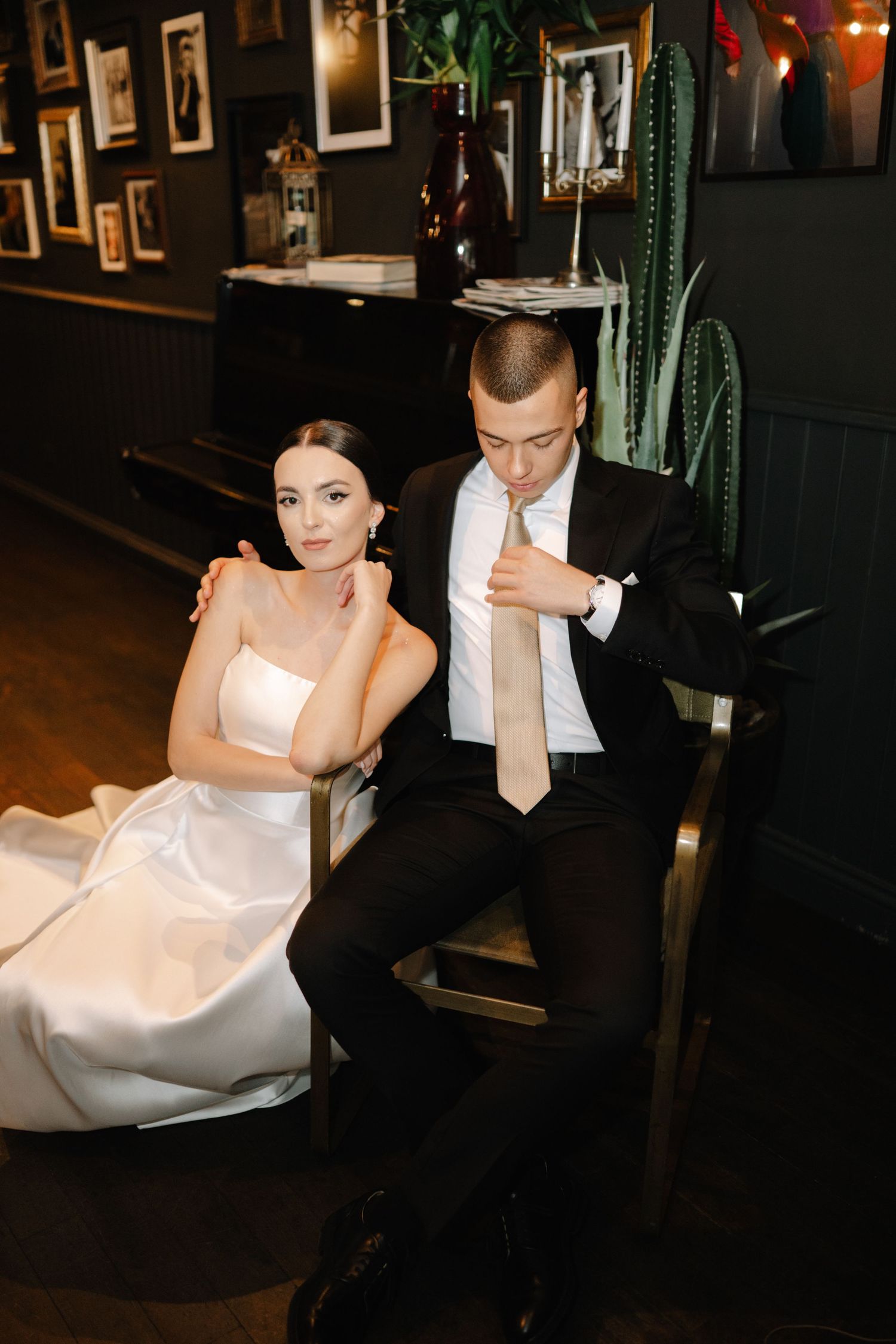 A bride and groom sit together in a dark room with framed photos on the wall behind them.