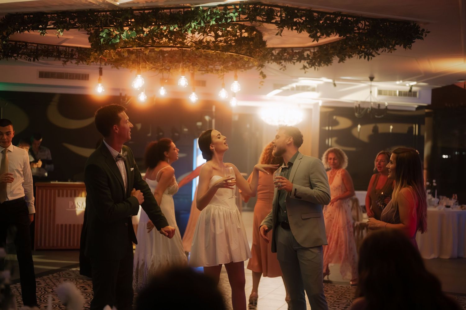 People dancing and celebrating at a dimly lit wedding reception with decorative lights and greenery overhead.