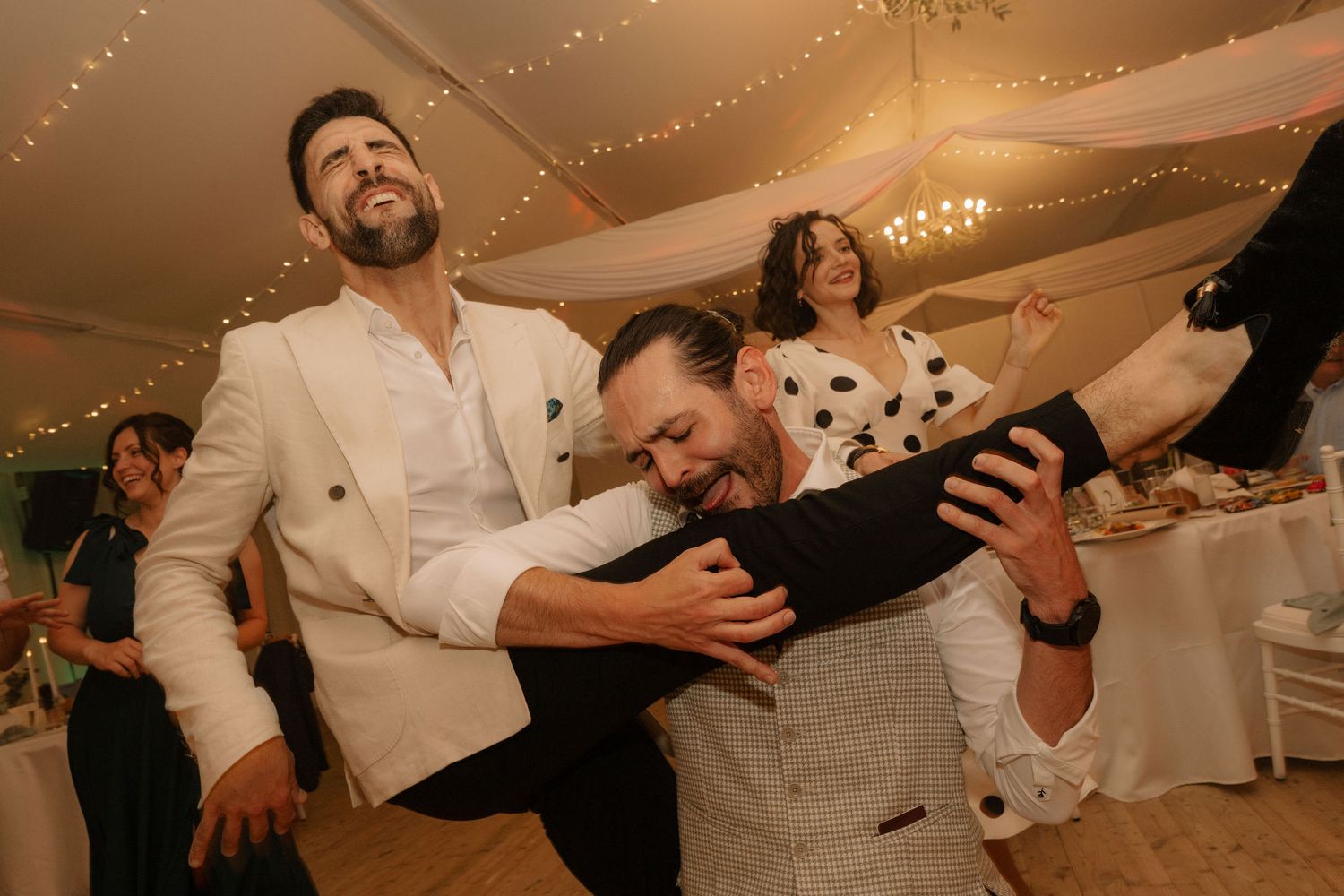 Two men dancing joyfully at a wedding reception in a marquee, with guests celebrating in the background.