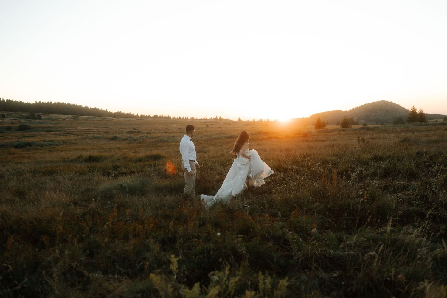 A bride and groom walk through a golden meadow at sunset with rolling hills in the background.