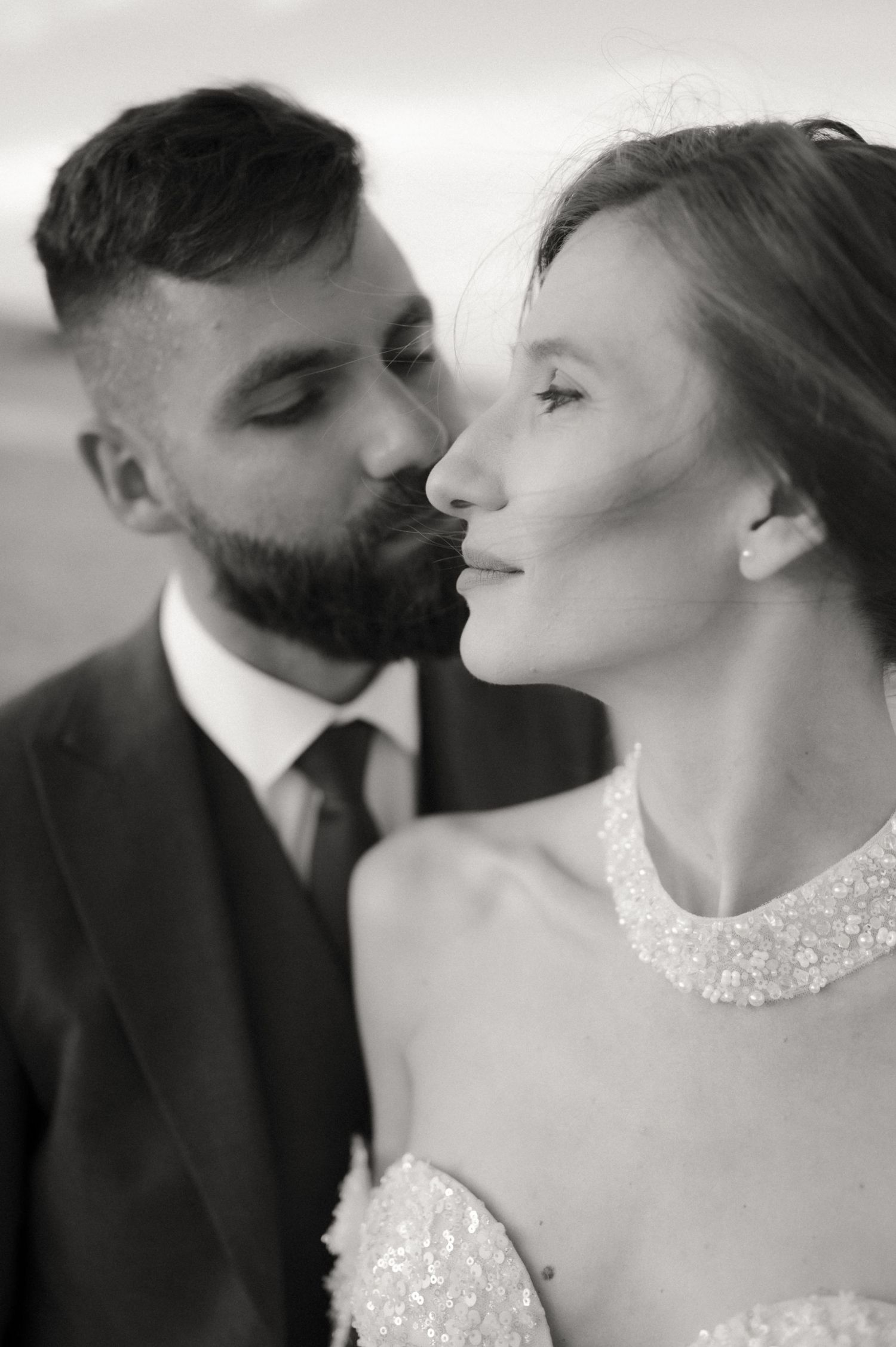 Black and white wedding photo of groom kissing bride on cheek, bride in embellished dress, groom in dark suit.