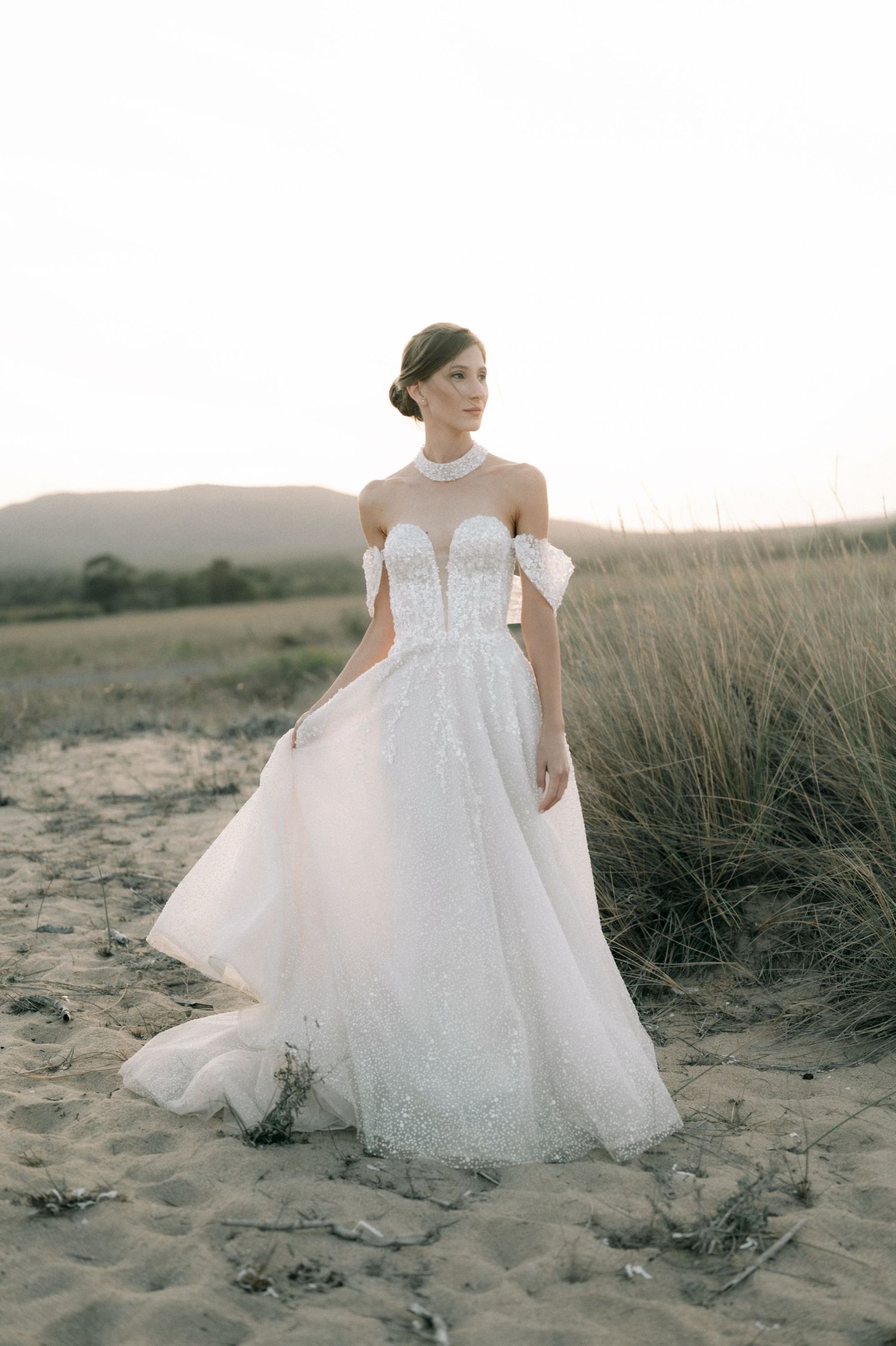 Bride in off-shoulder lace ball gown wedding dress standing on sandy beach with golden grass backdrop.