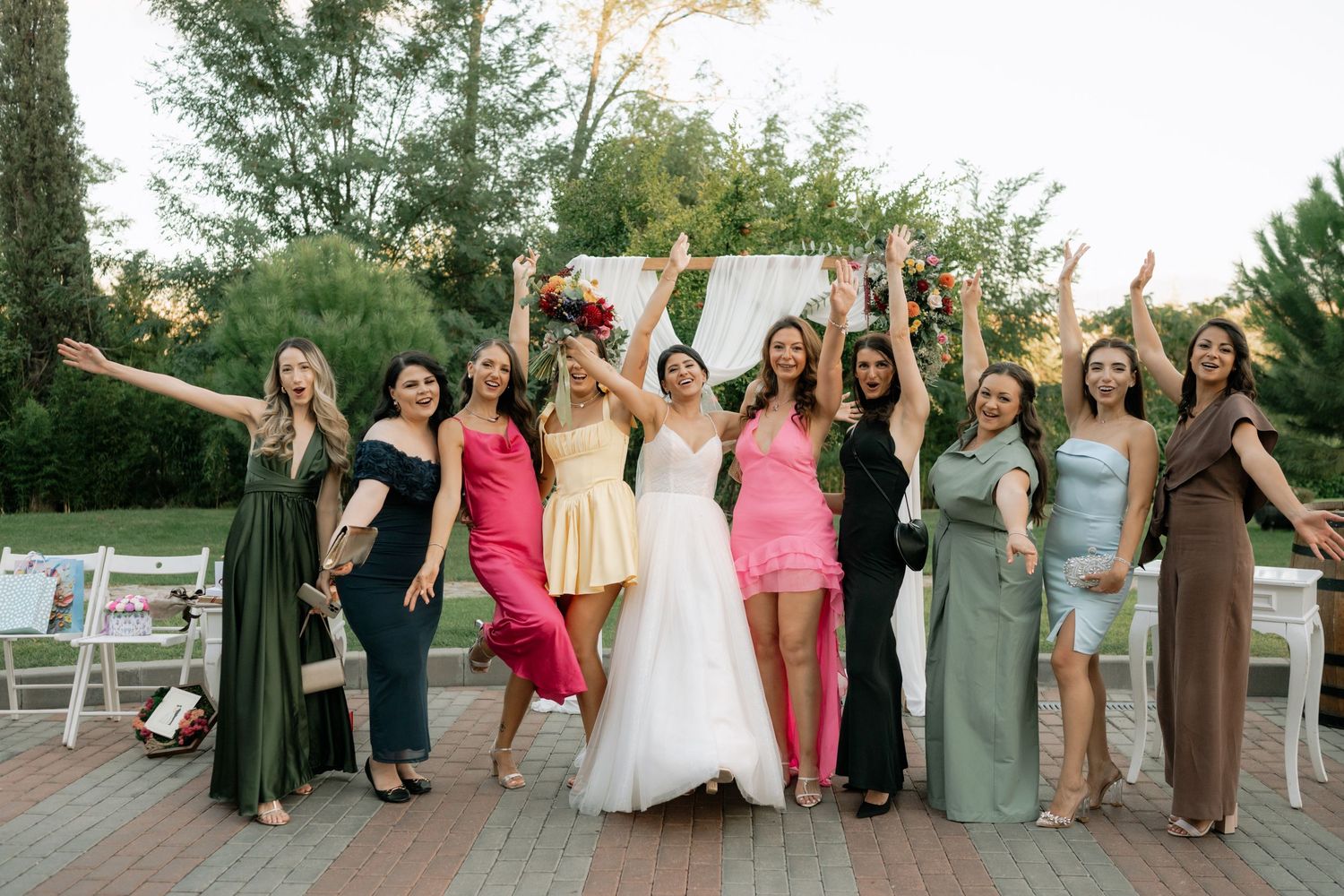 A group of people in formal dresses pose together outdoors with raised arms in celebration at an event.