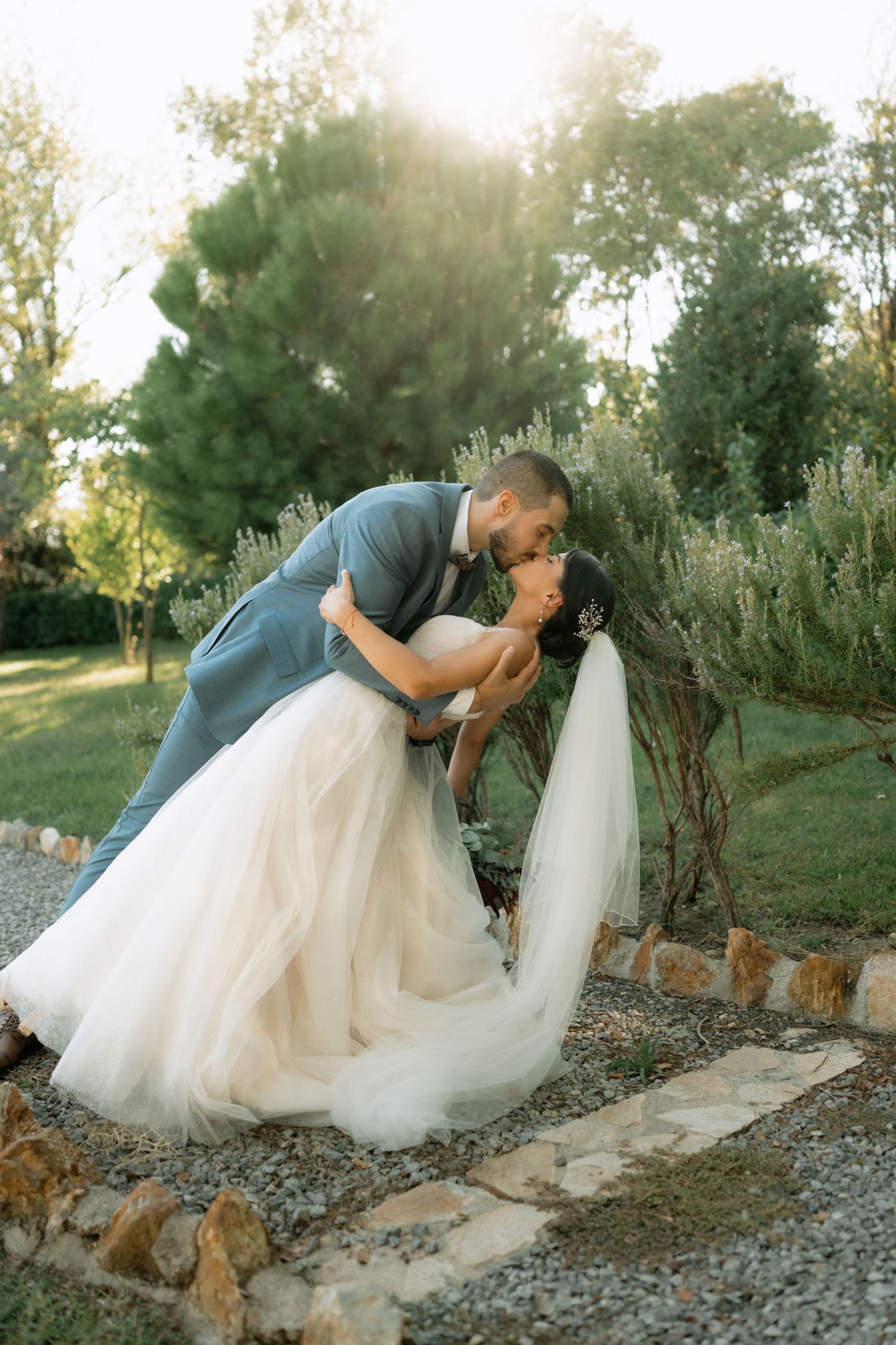 A romantic wedding couple embraces in a garden setting during golden hour, with the bride wearing a flowing tulle gown.
