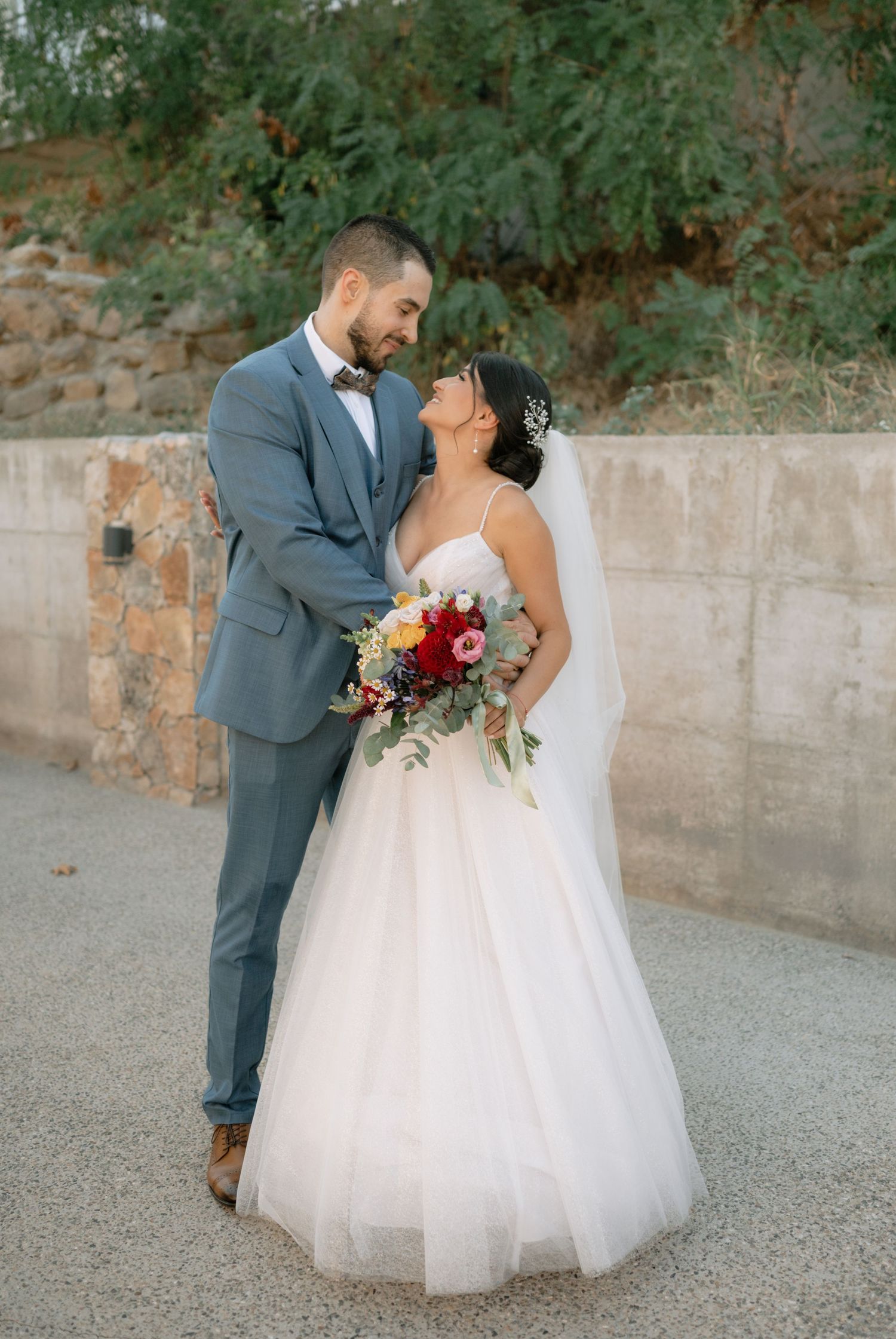 A romantic wedding couple shares intimate moments on a gravel path against a stone wall and trees in the background.