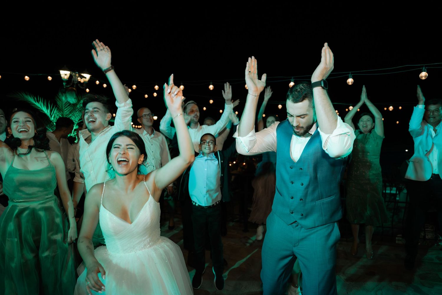 Wedding guests dance together on a dark dance floor illuminated by party lights during an evening reception celebration.