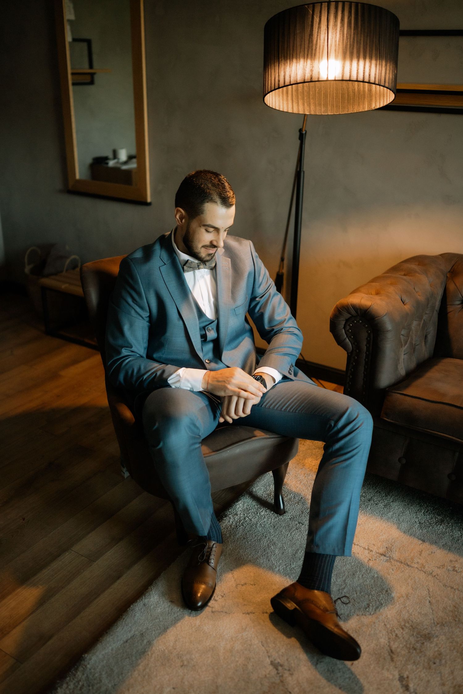 A person in a blue suit sits contemplatively on a gray couch in a dimly lit room with pendant lighting above.