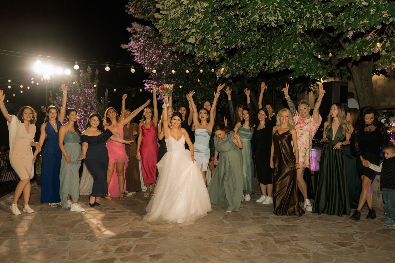 Wedding guests and bride celebrate with arms raised during a nighttime outdoor reception.