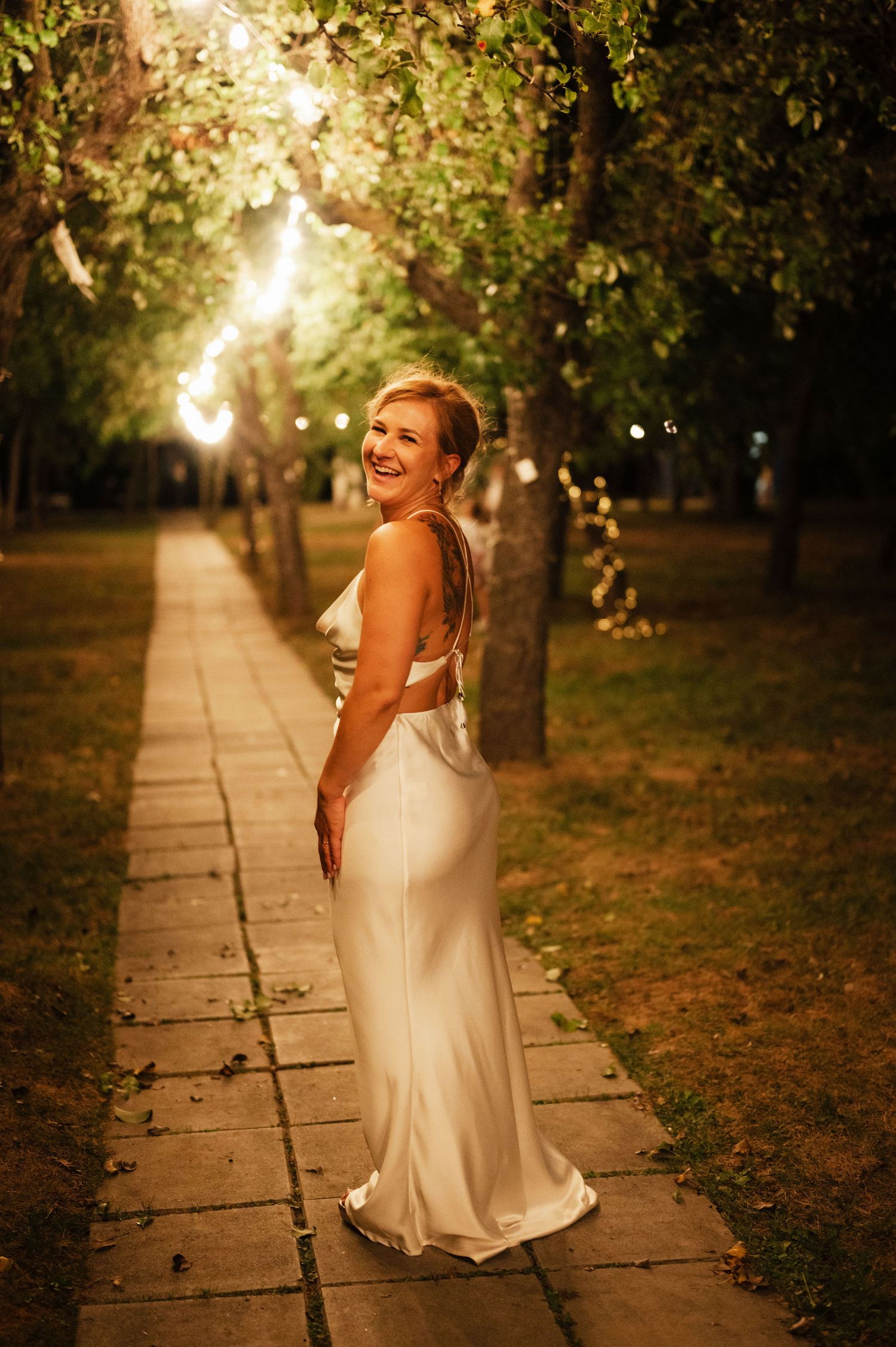 A bride in a flowing white dress twirls joyfully on a stone path surrounded by trees at golden hour.