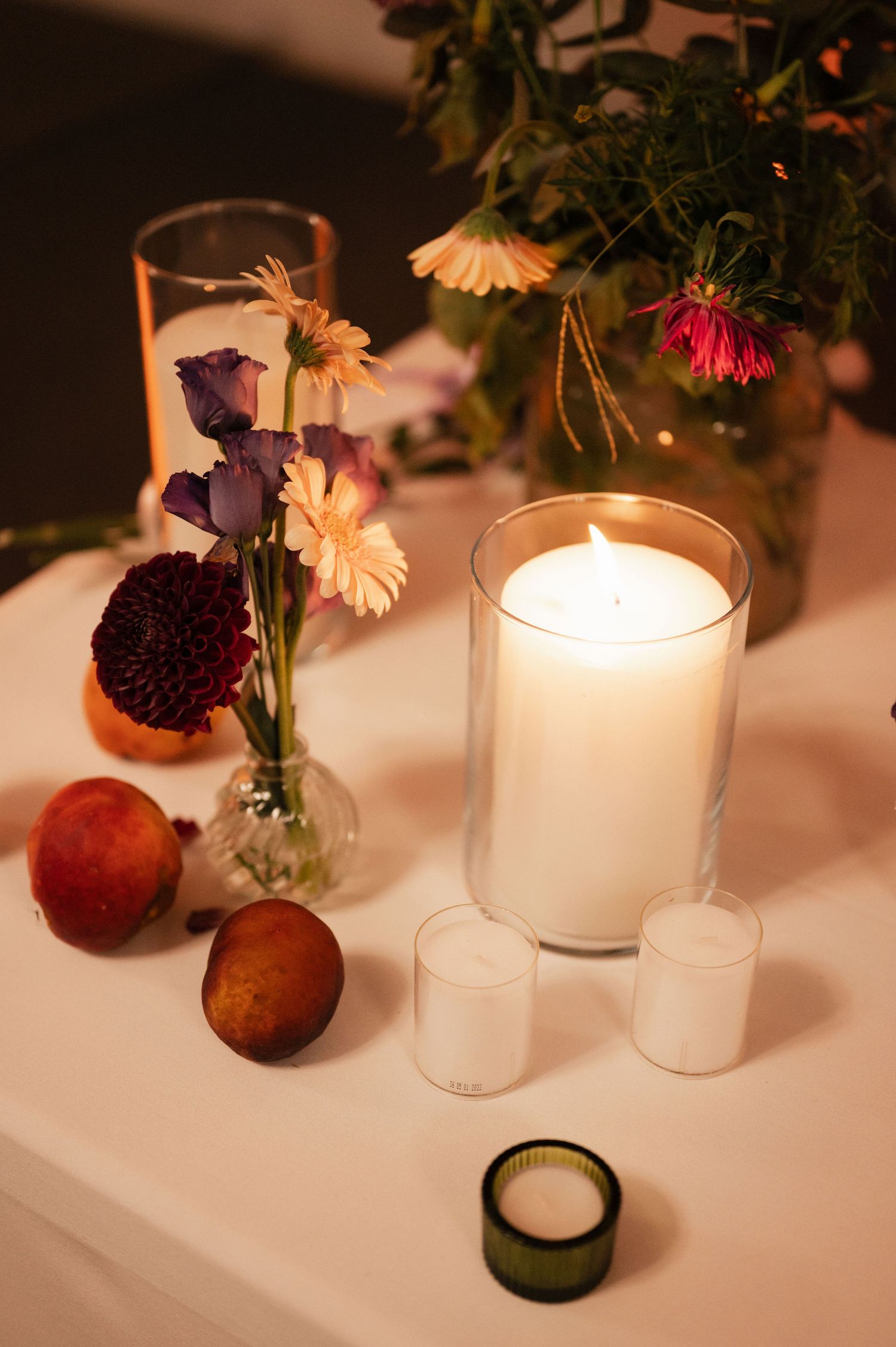 Elegant table setting with glowing white candles and small floral arrangements in rustic vases against a dark background.