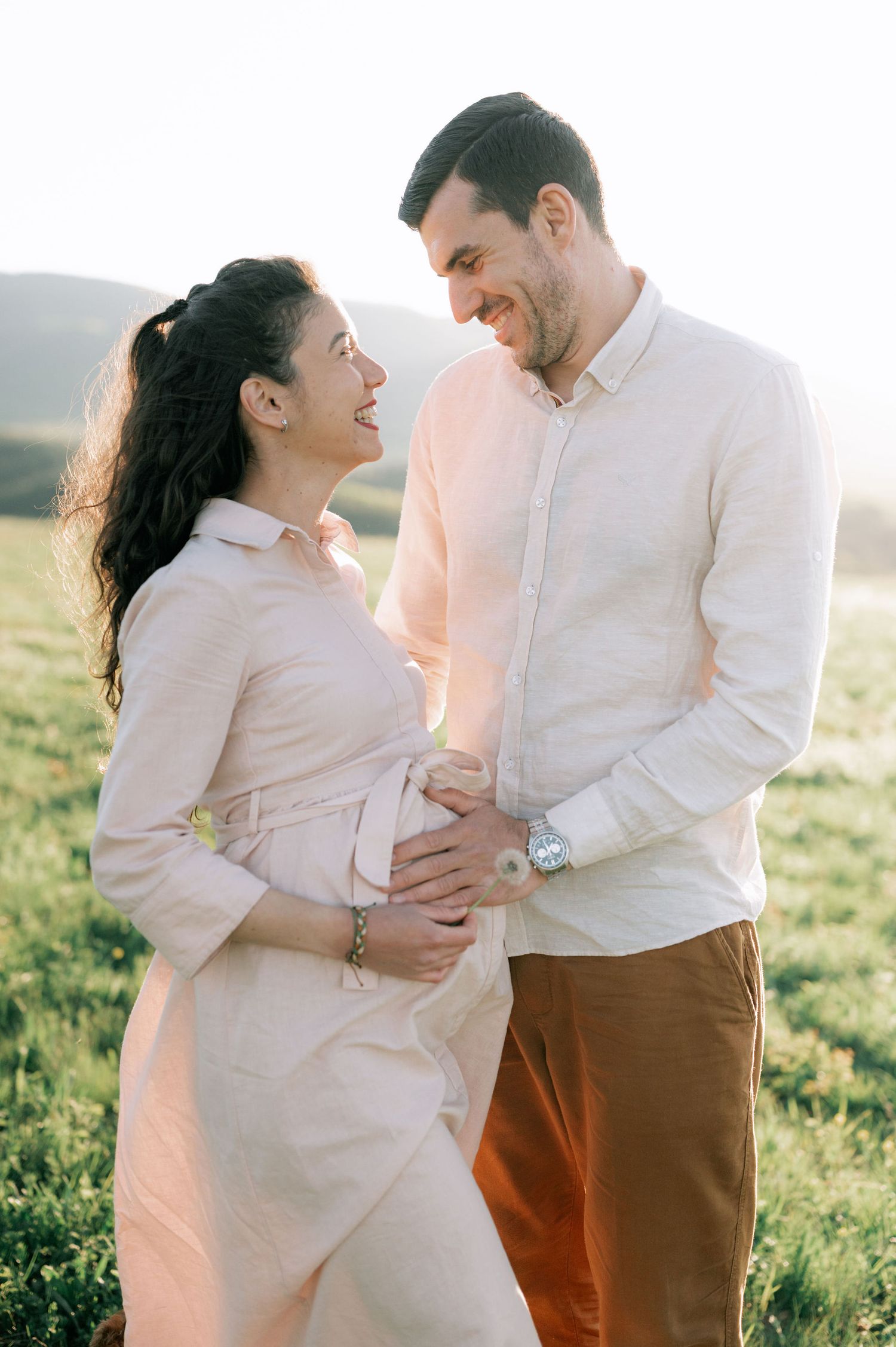 A couple in light colored clothing share a tender moment in a sunlit field during a maternity photoshoot.