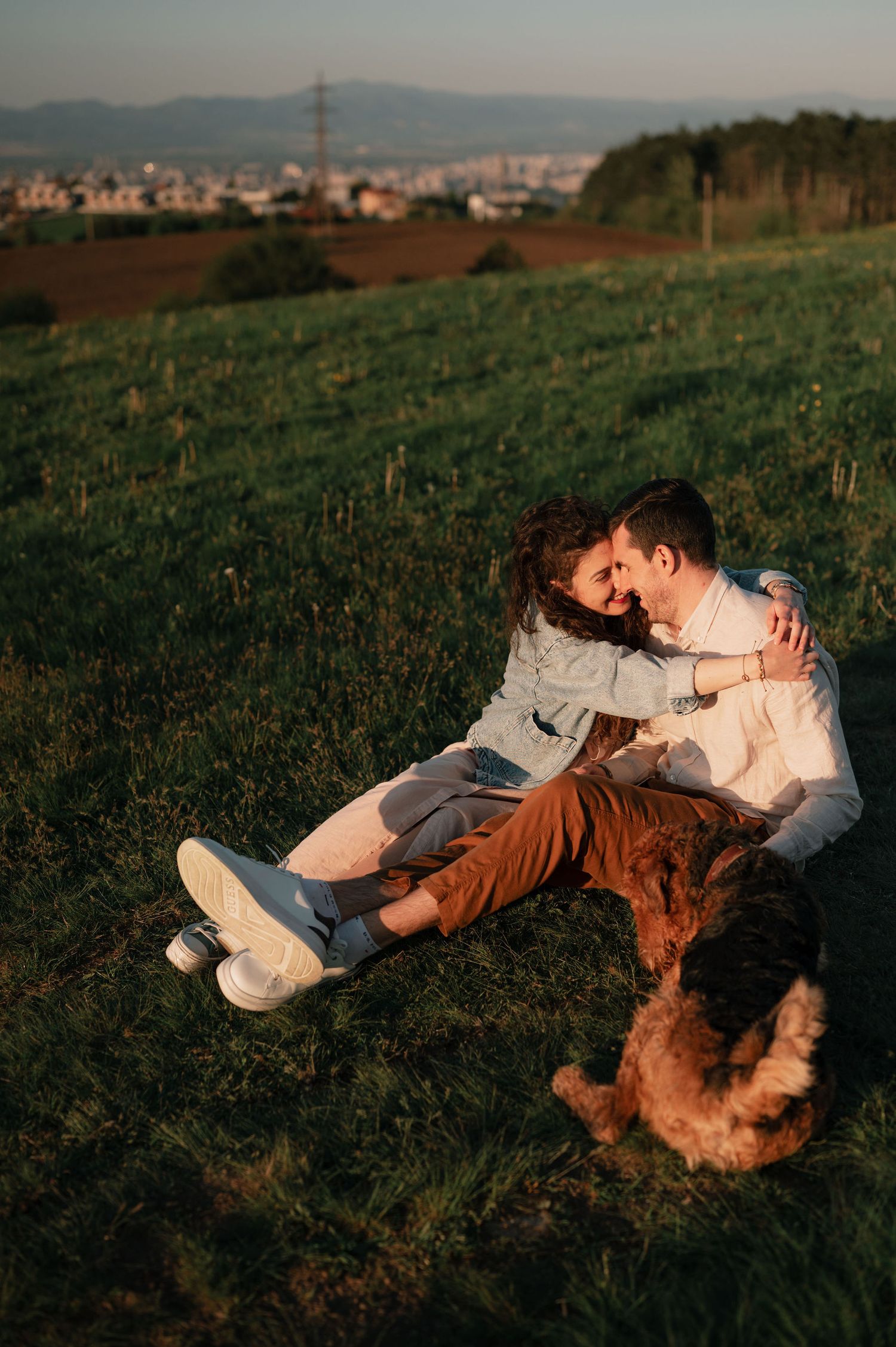 Couple cuddles on grassy hill with their dog while overlooking distant cityscape.