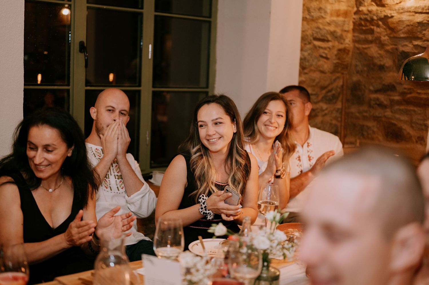 Wedding guests laugh and celebrate during an intimate indoor reception with exposed stone walls and warm lighting.