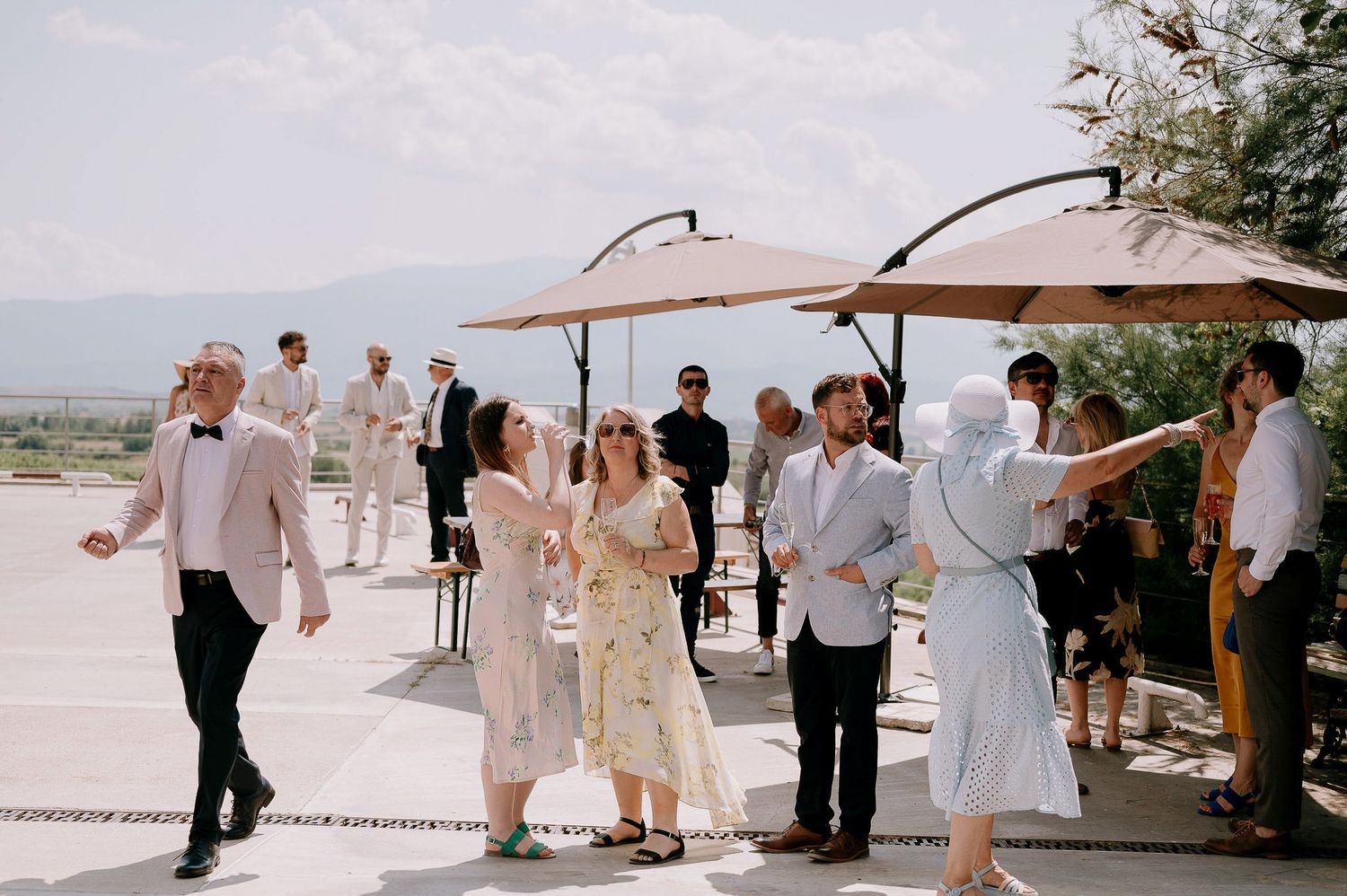 Wedding guests gather under umbrellas at an outdoor celebration on a sunny day.