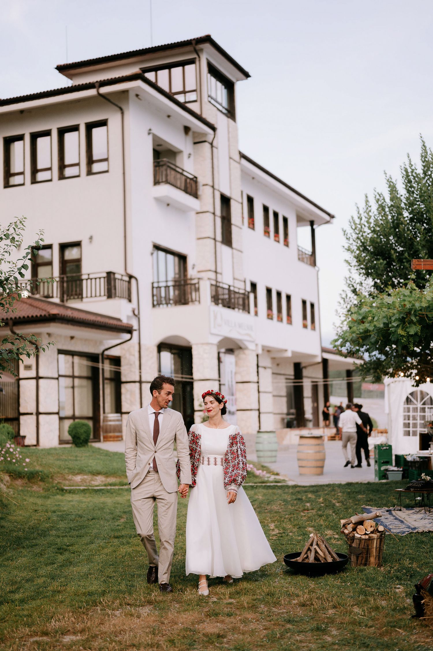 Elegant outdoor celebration at a white villa with manicured lawn and bonfire setup.