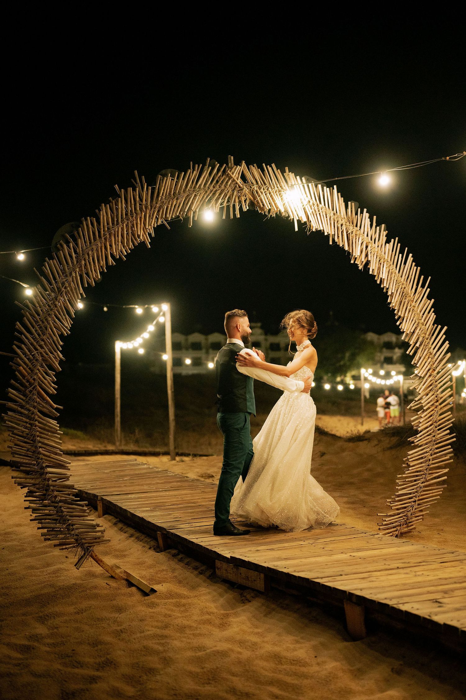 Romantic wooden boardwalk at night with illuminated driftwood arch and string lights.
