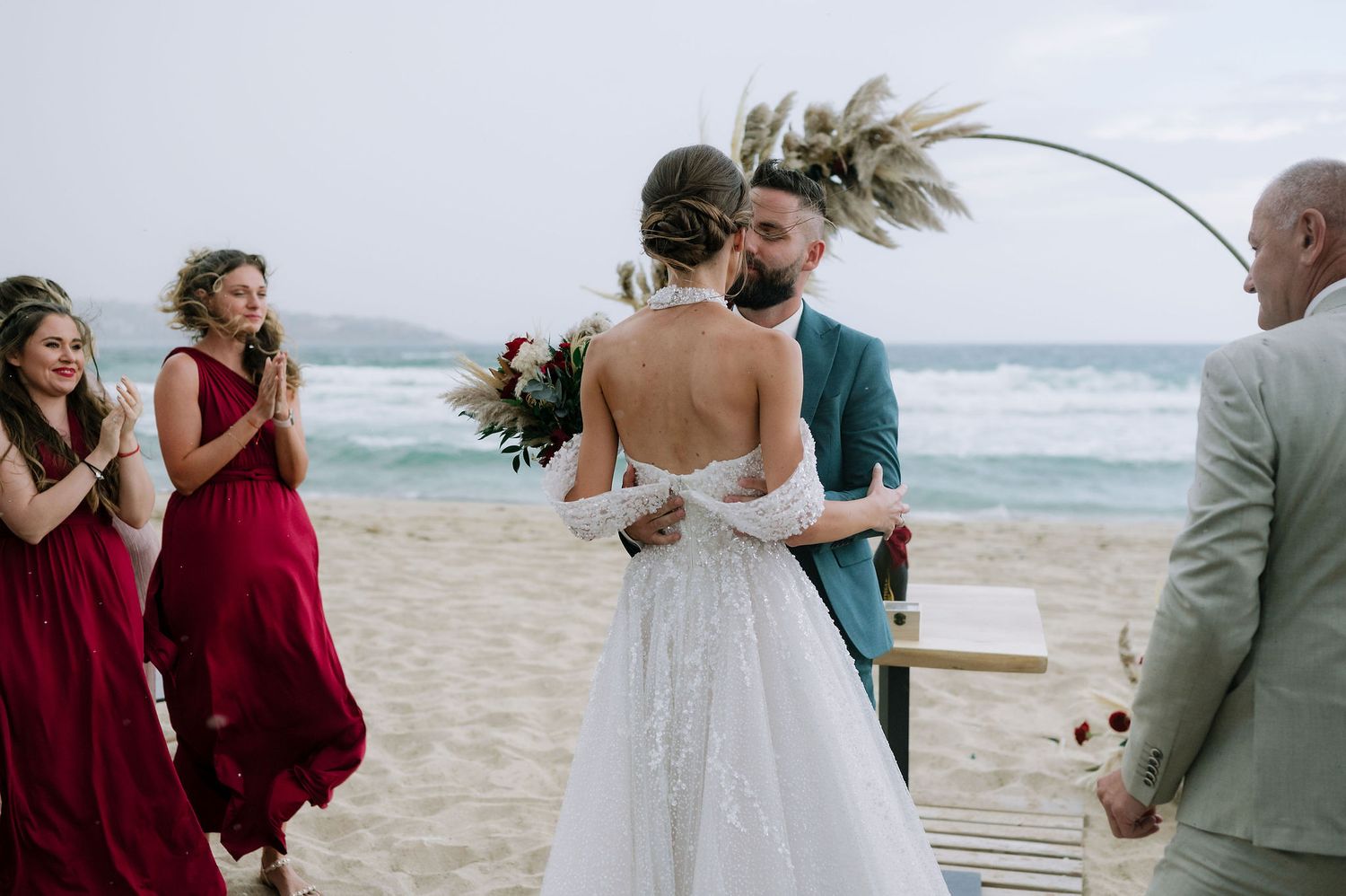 Wedding celebration on a windy beach with guests gathered around a wooden walkway.