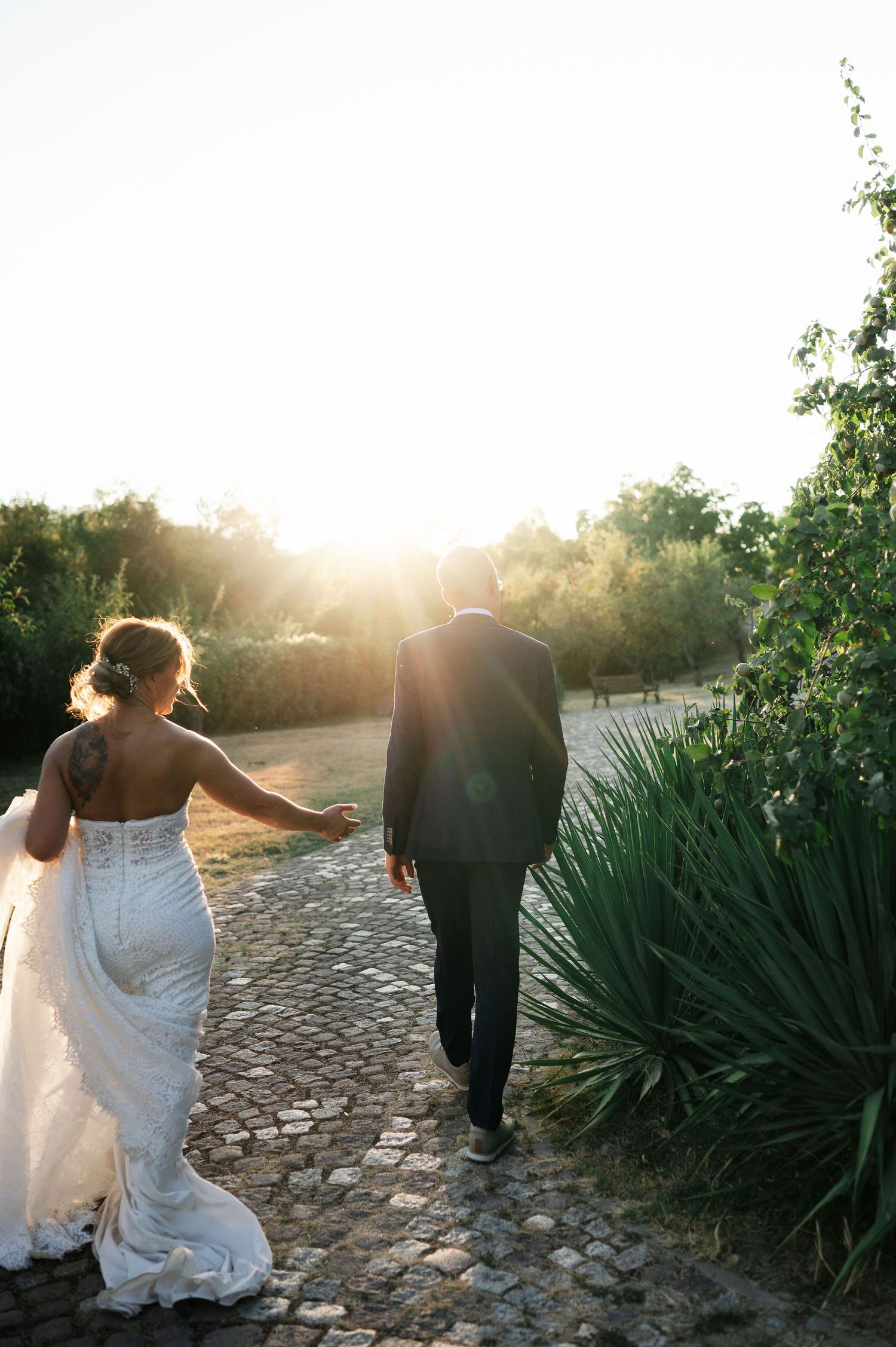 A couple walking on a cobblestone path at sunset with the bride's white dress trailing behind.