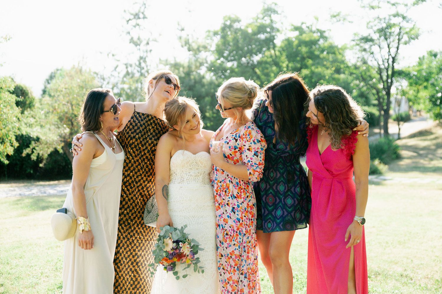 A bridal party in mixed floral and solid dresses poses outdoors on a sunny day.