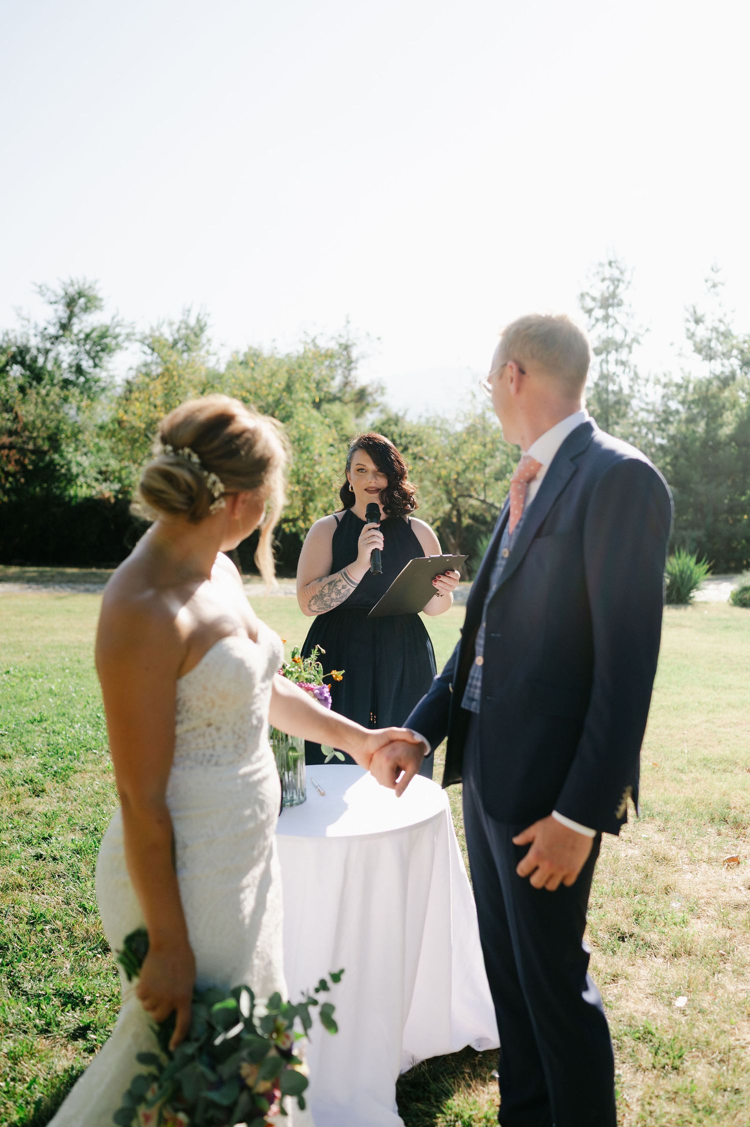 A wedding ceremony takes place in a sunny outdoor field with guests gathered for the special moment.