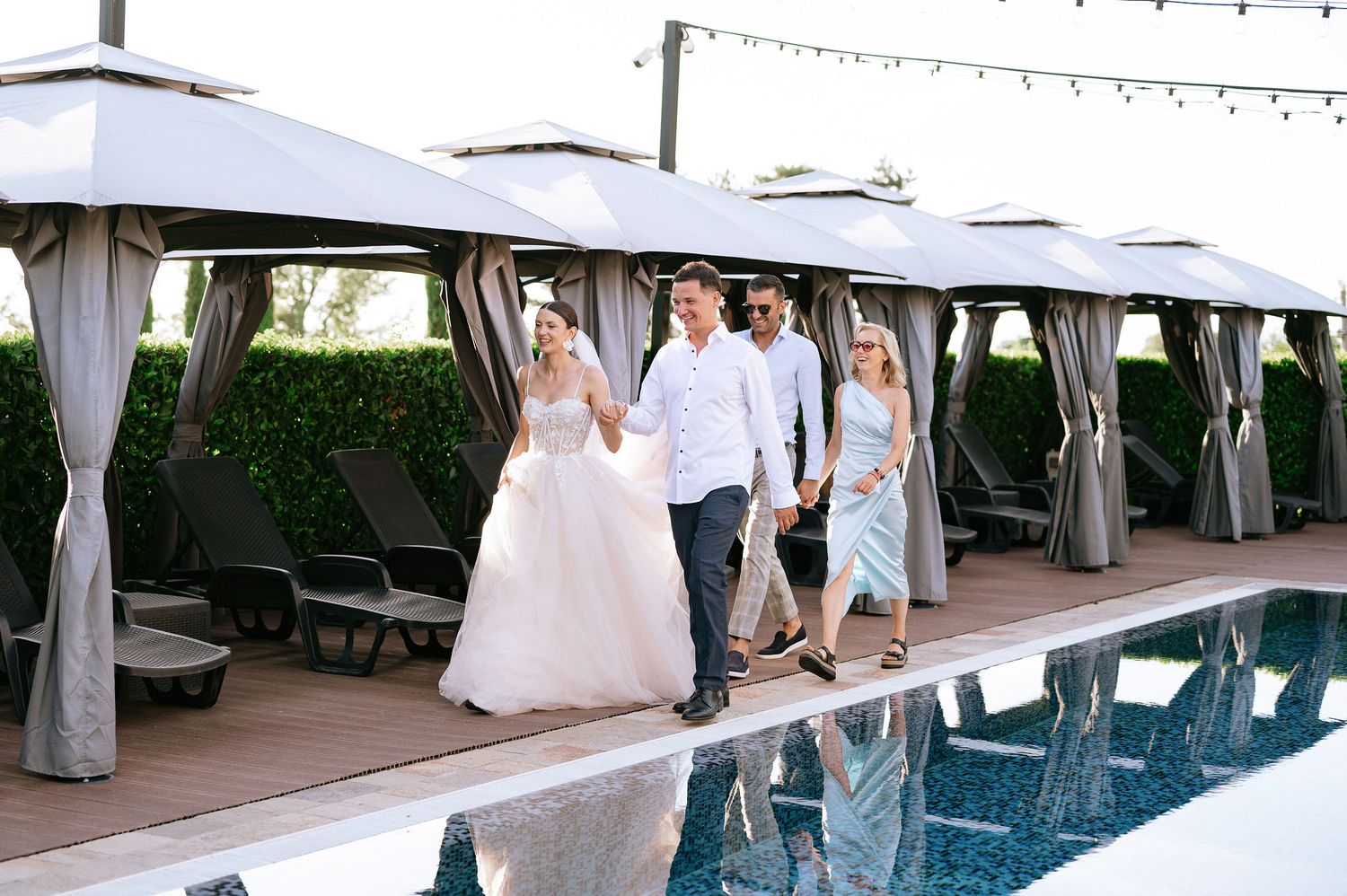 Wedding party walks alongside a swimming pool with cabanas and string lights.