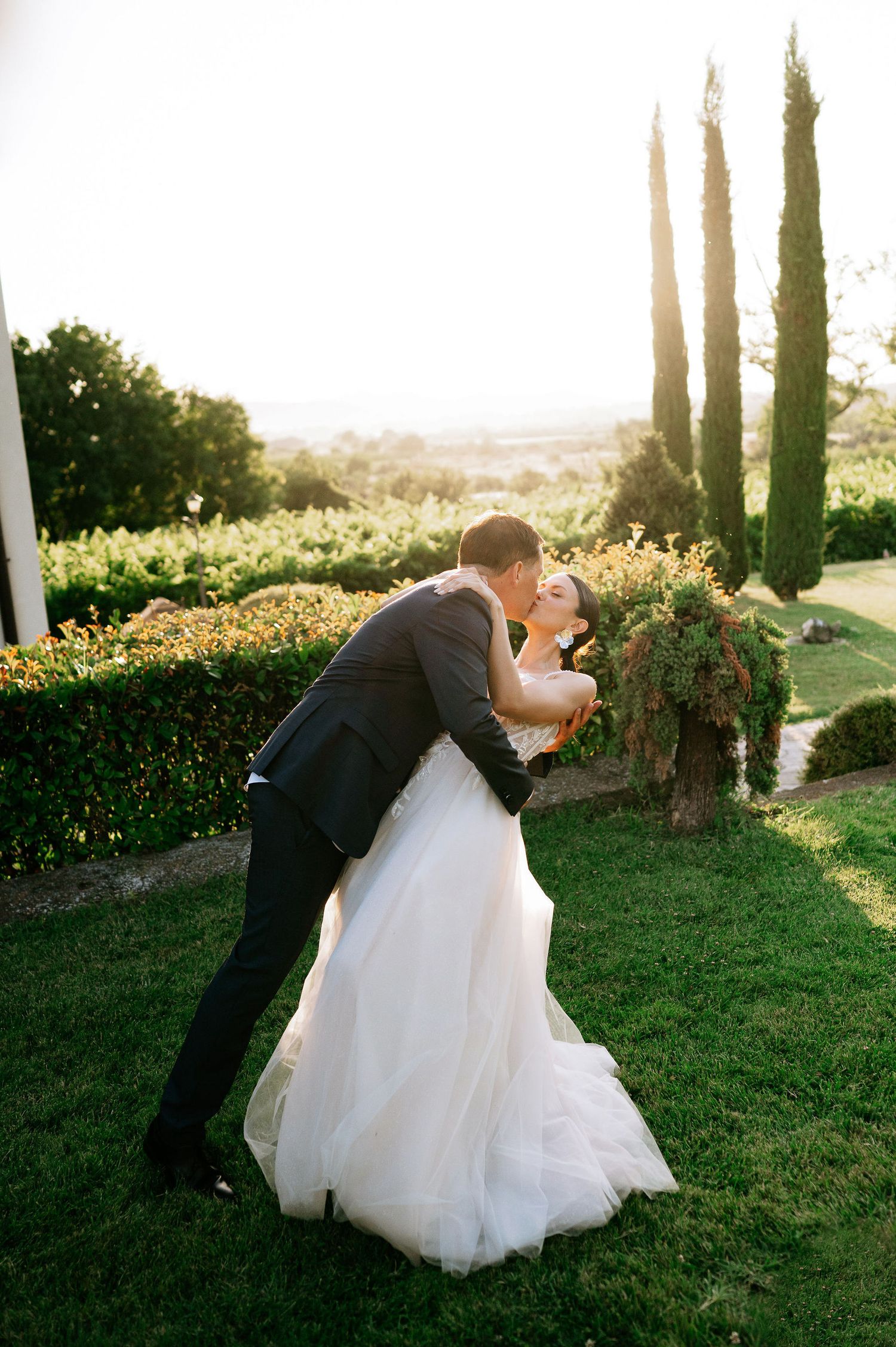 Romantic dip pose between newlyweds on a grassy hillside with cypress trees.