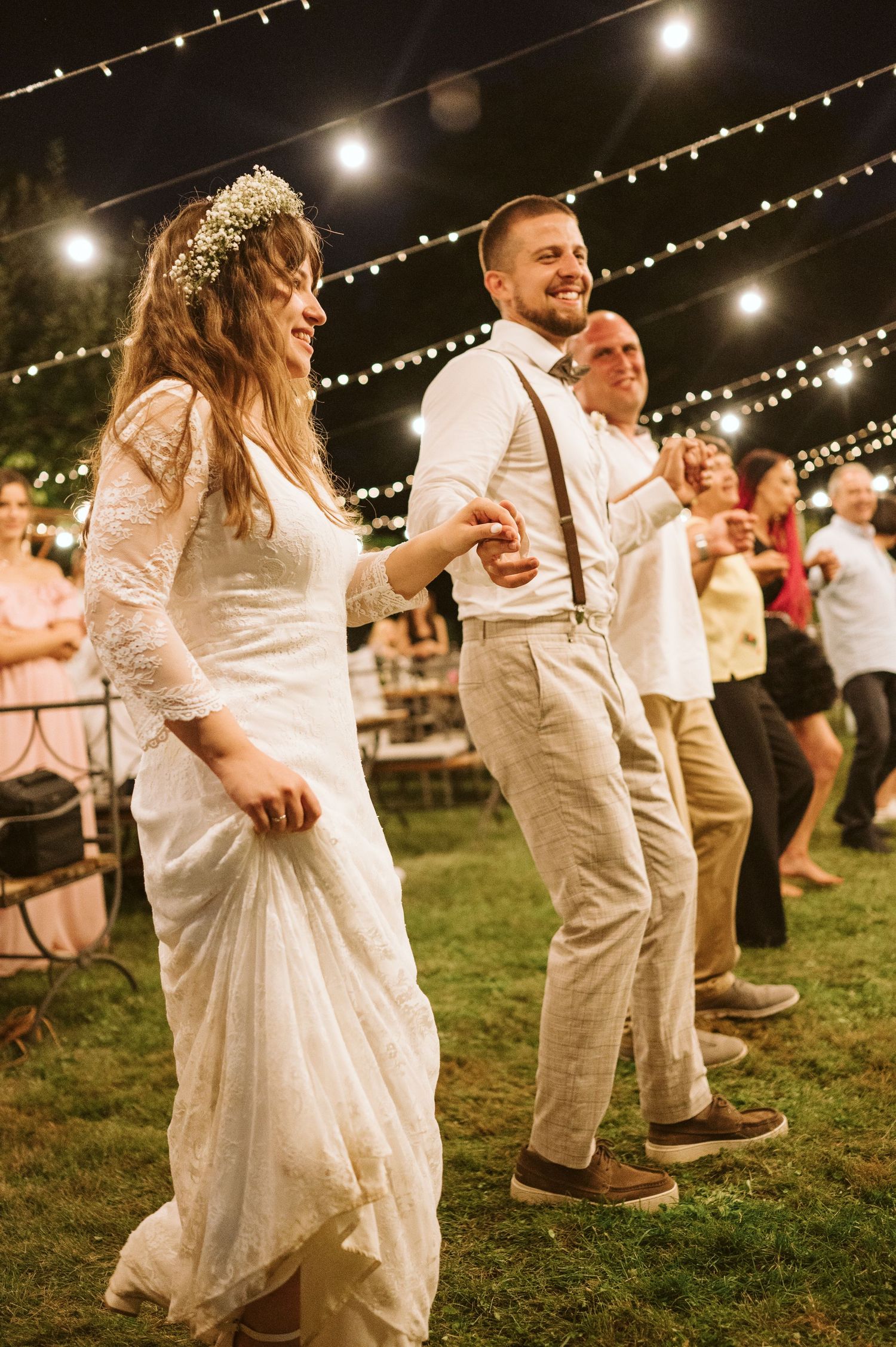 Wedding guests dance under twinkling string lights at an outdoor evening reception.