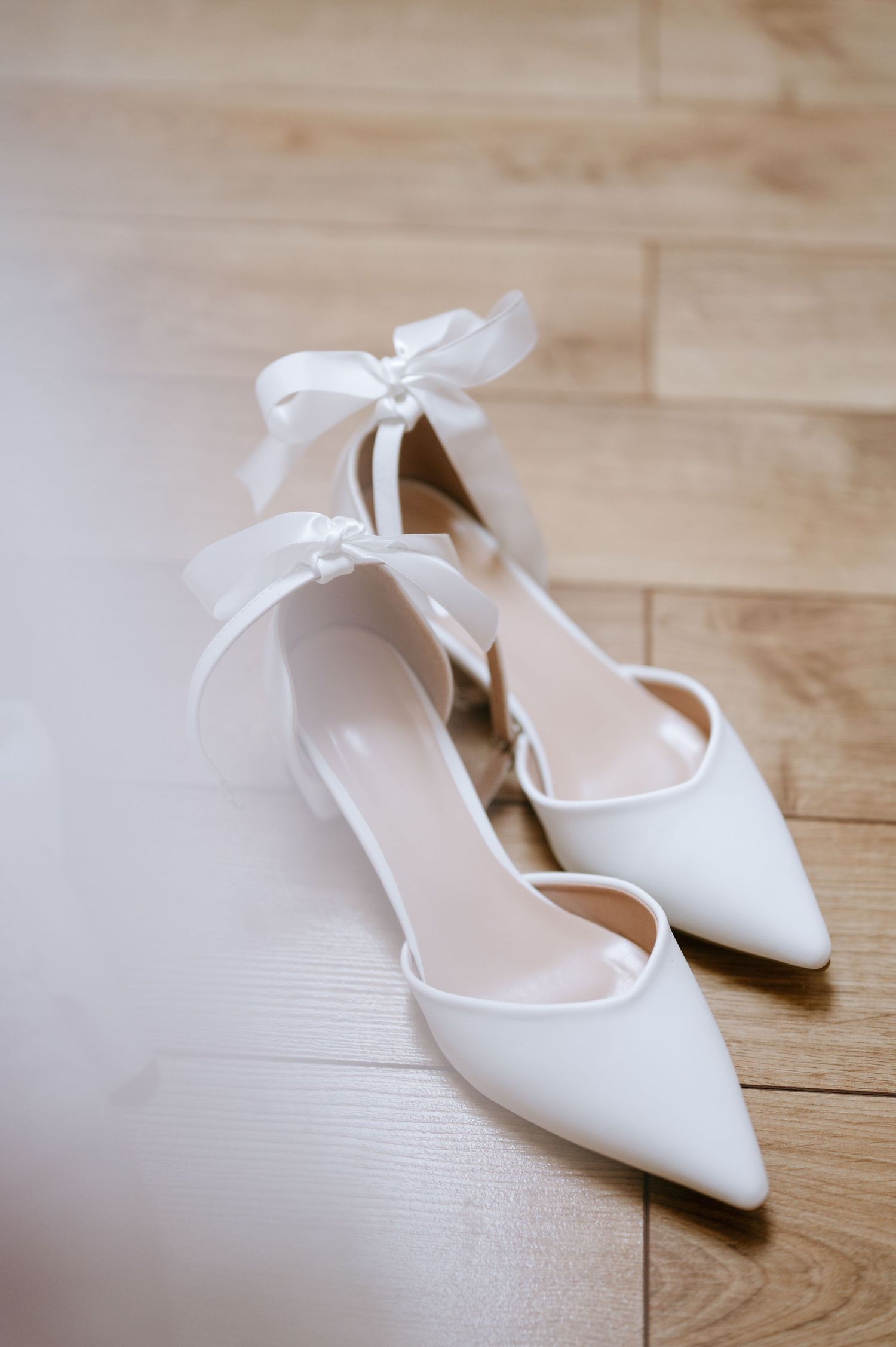 Elegant white bridal shoes with delicate bow details rest on a wooden floor.