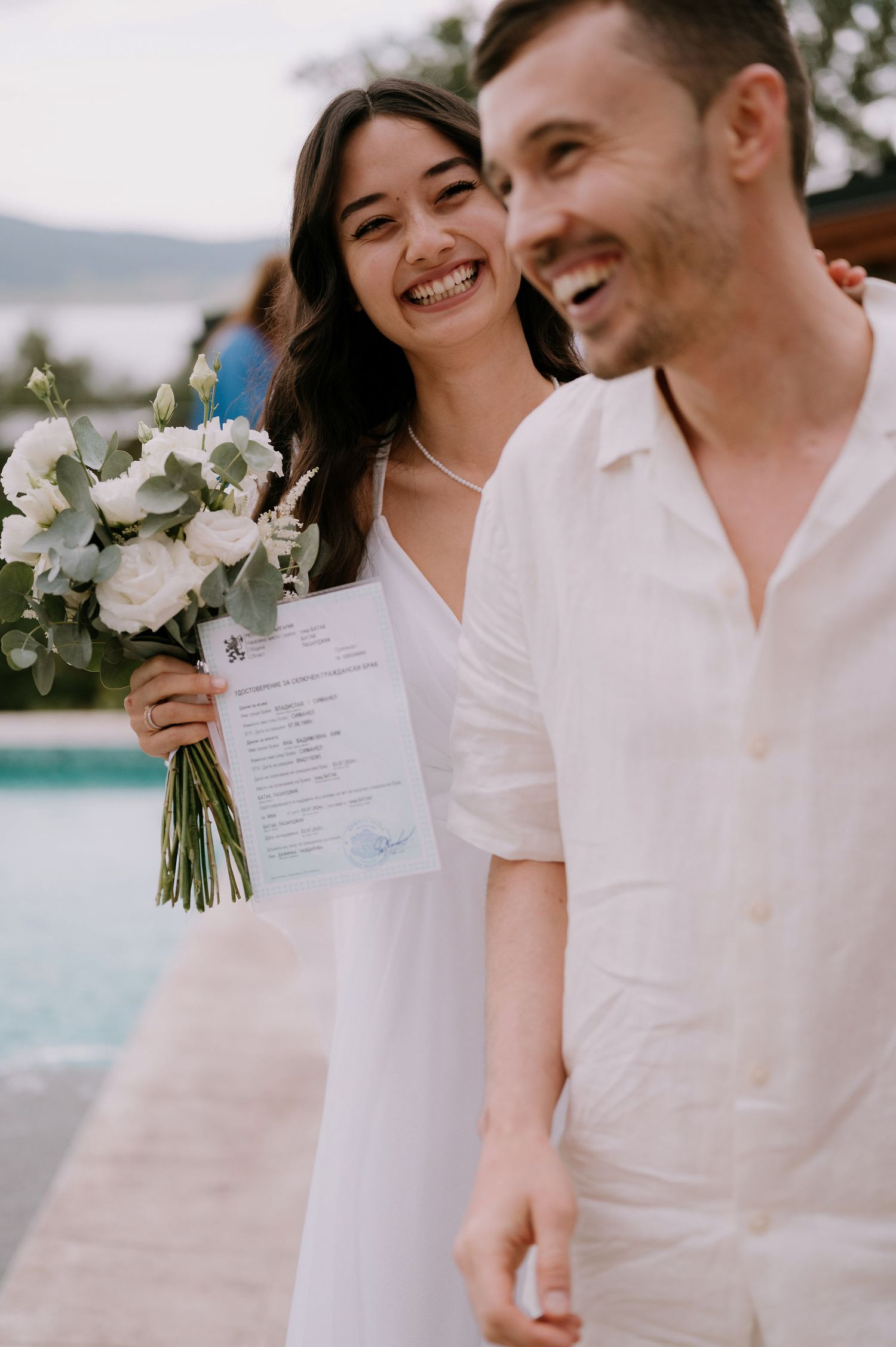 A couple in white attire shares joyful moments by the pool during their wedding celebration.