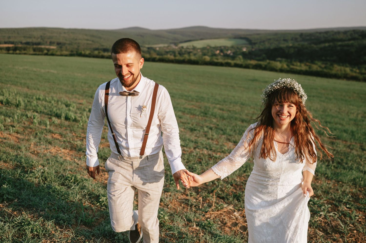 Two people in white wedding clothes dance and laugh together on a grassy hillside.