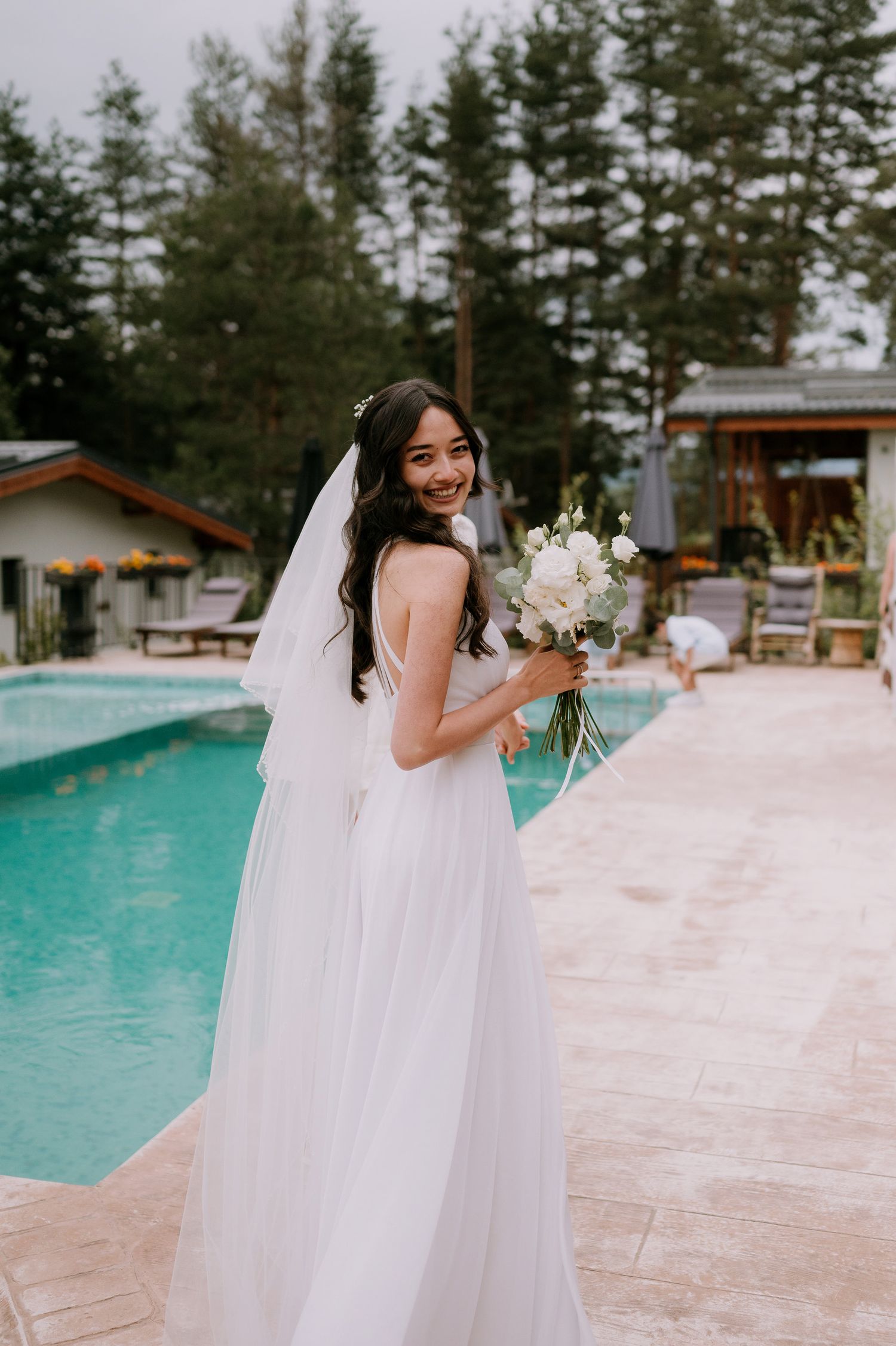 A bride in a flowing white dress poses gracefully beside a swimming pool surrounded by evergreen trees.