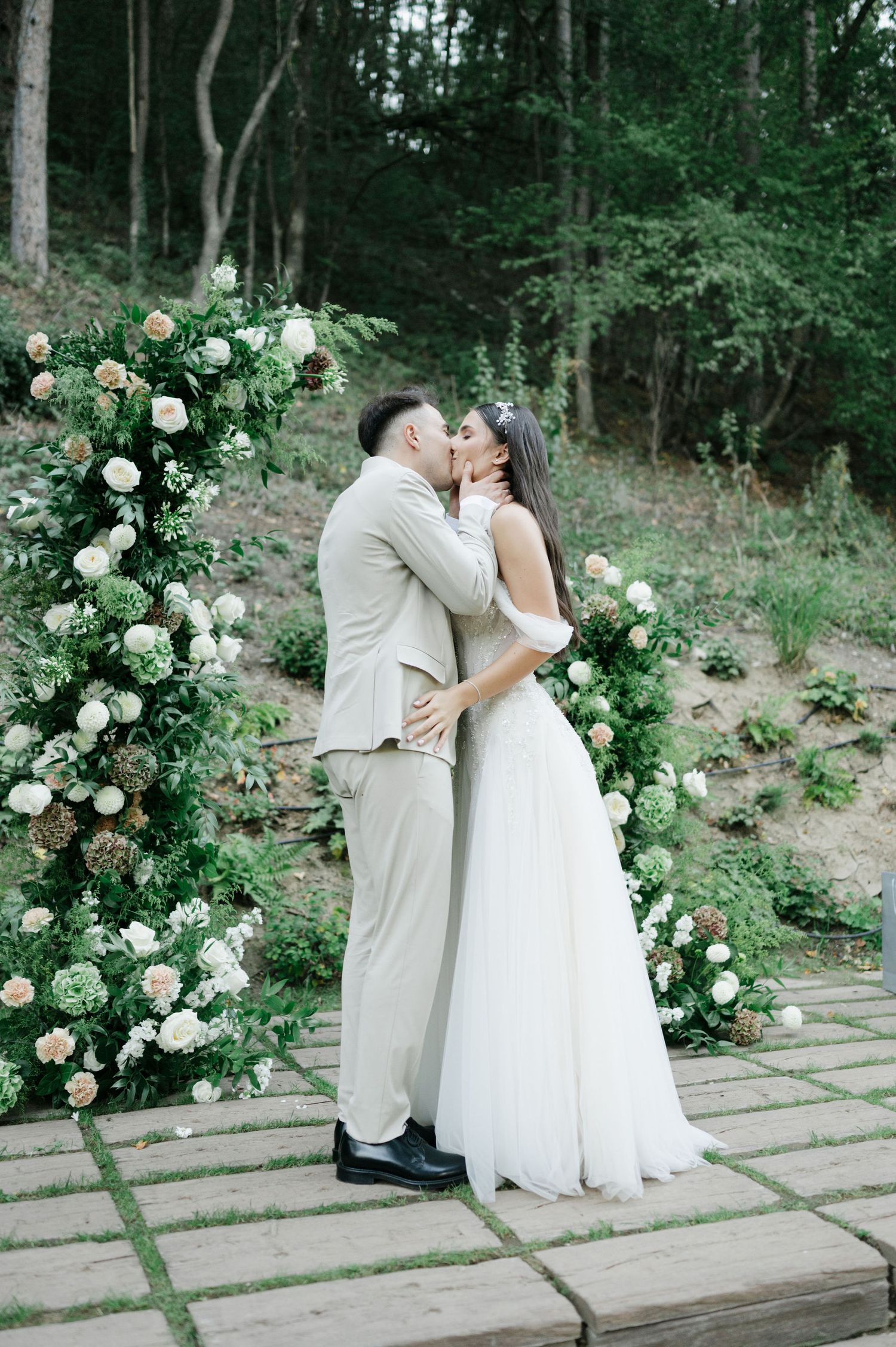 Couple embraces during their woodland wedding ceremony beside elegant white floral arrangements.