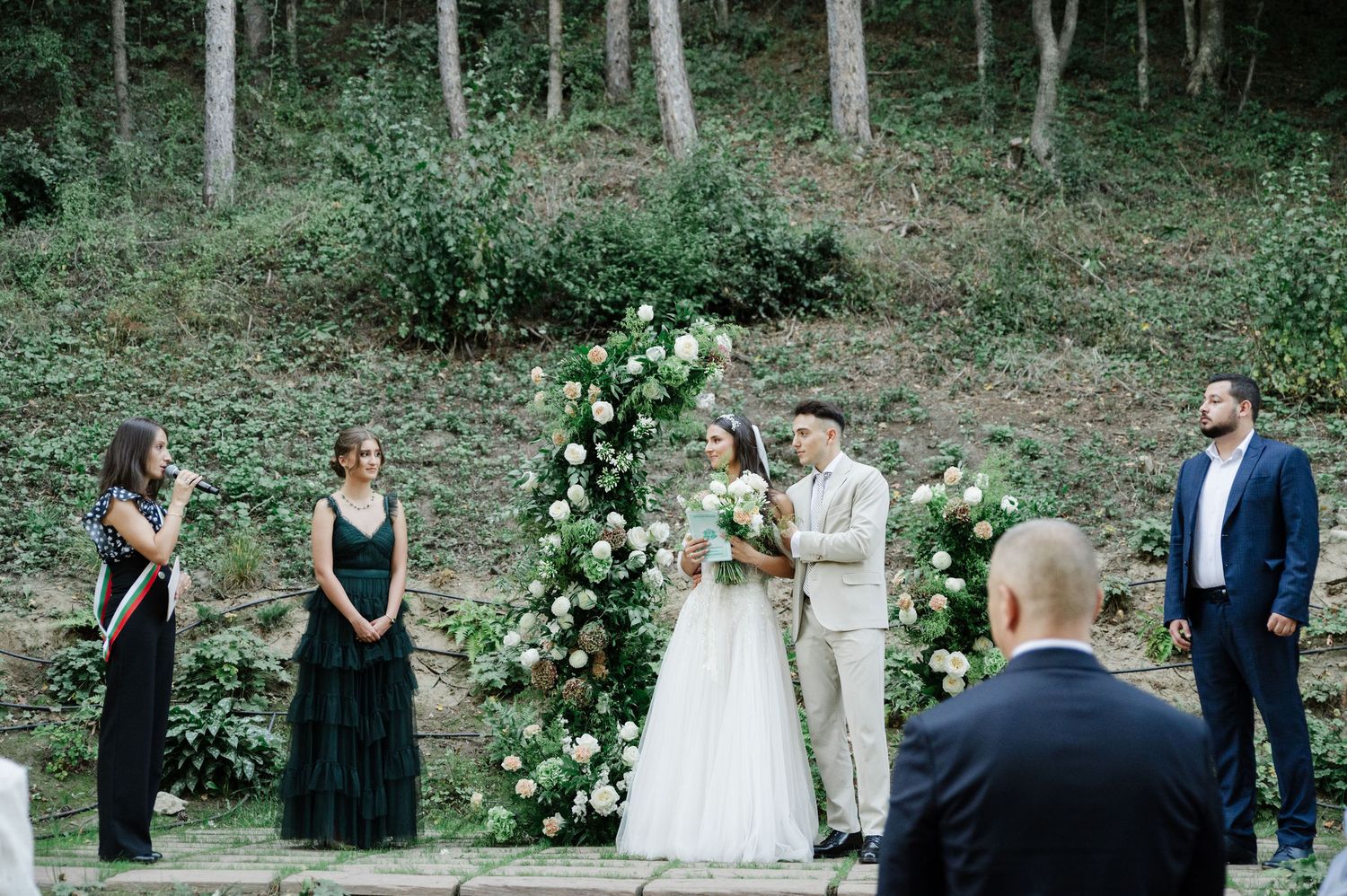 Outdoor forest wedding ceremony with guests seated on wooden benches surrounded by trees.