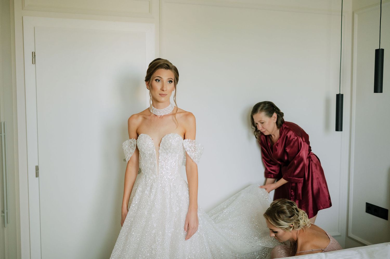 Wedding preparation scene with dress adjustments in a white room.