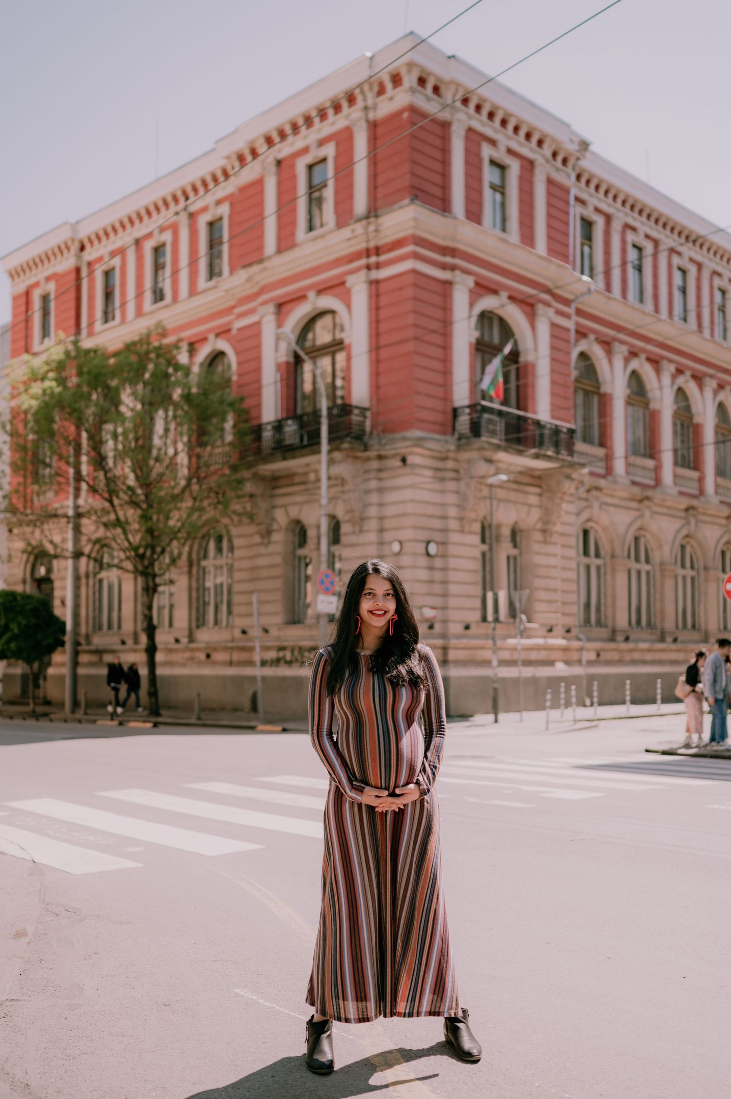 Woman in striped maxi dress smiling in front of a red historic building on a sunny day.