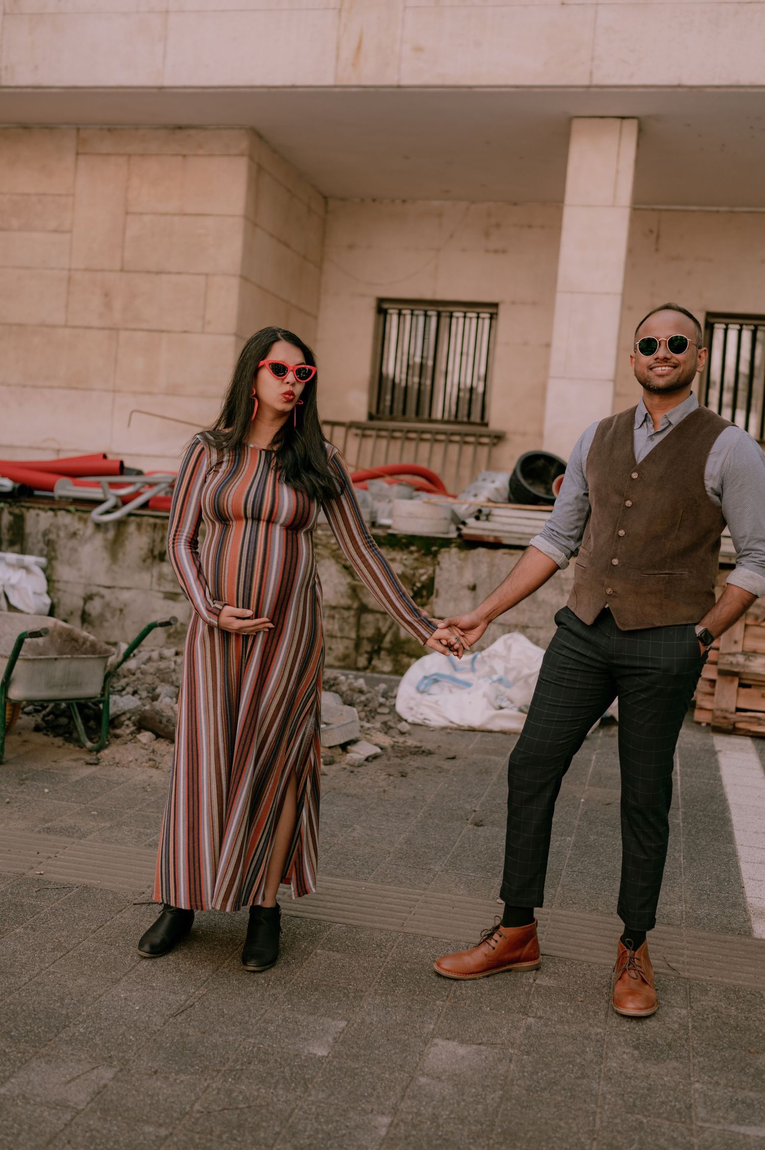 Pregnant woman in striped dress holds hands with smiling man in vest and sunglasses outdoors.
